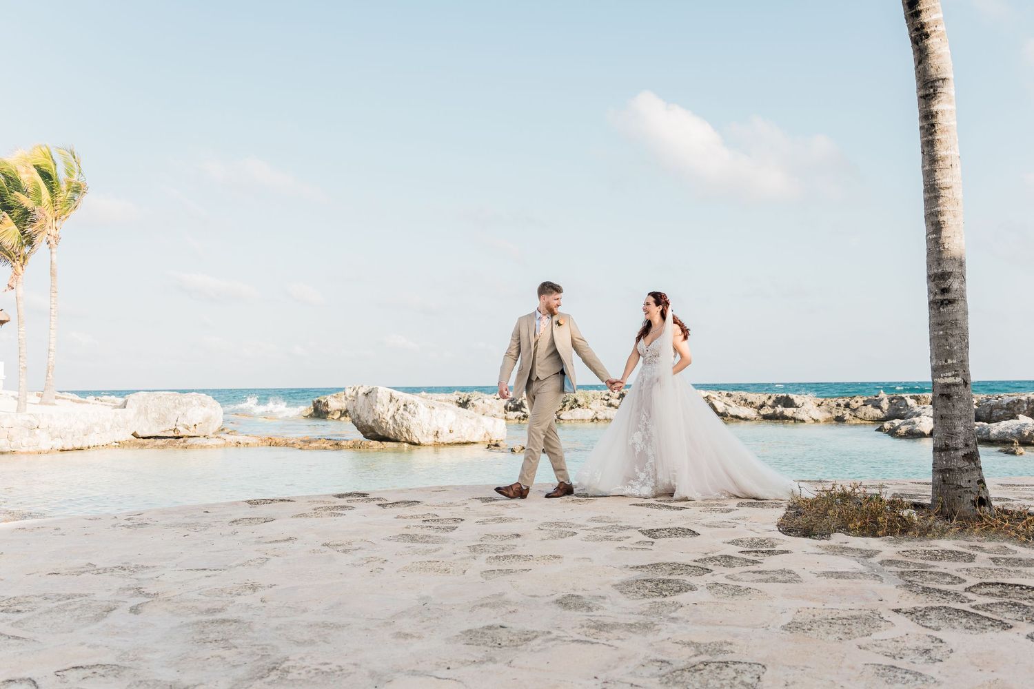 Bride and groom walk hand in hand along a rocky Caribbean beach near a palm tree.