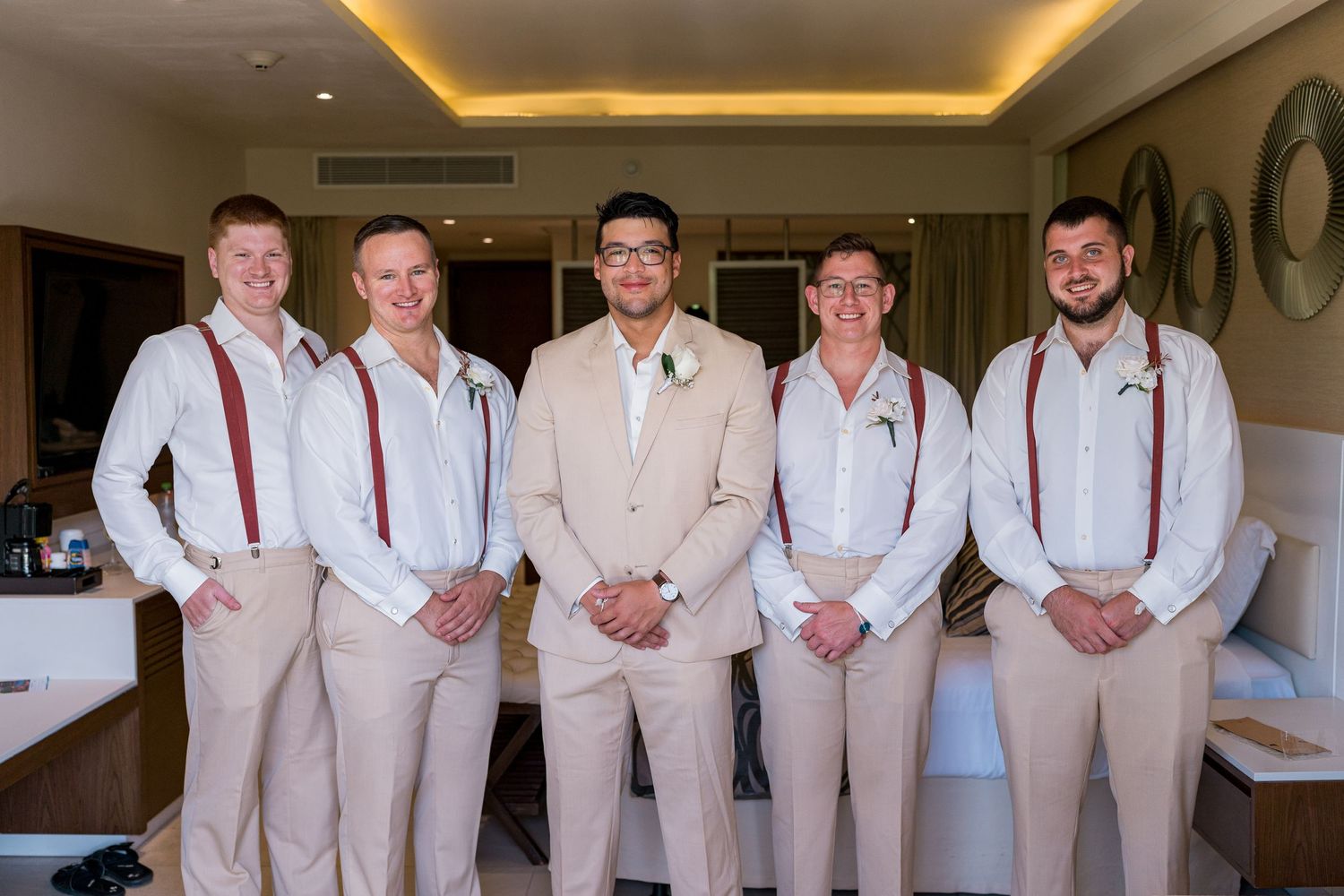 Groom and four groomsmen in beige suits and red suspenders posing together in a hotel room.