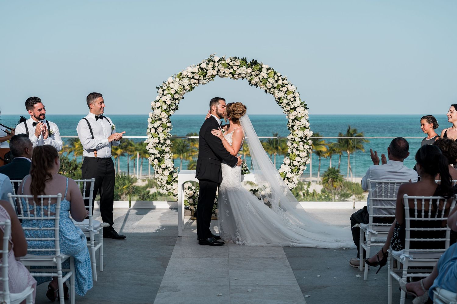 Newlyweds share first kiss under white floral arch at stunning oceanfront wedding ceremony.