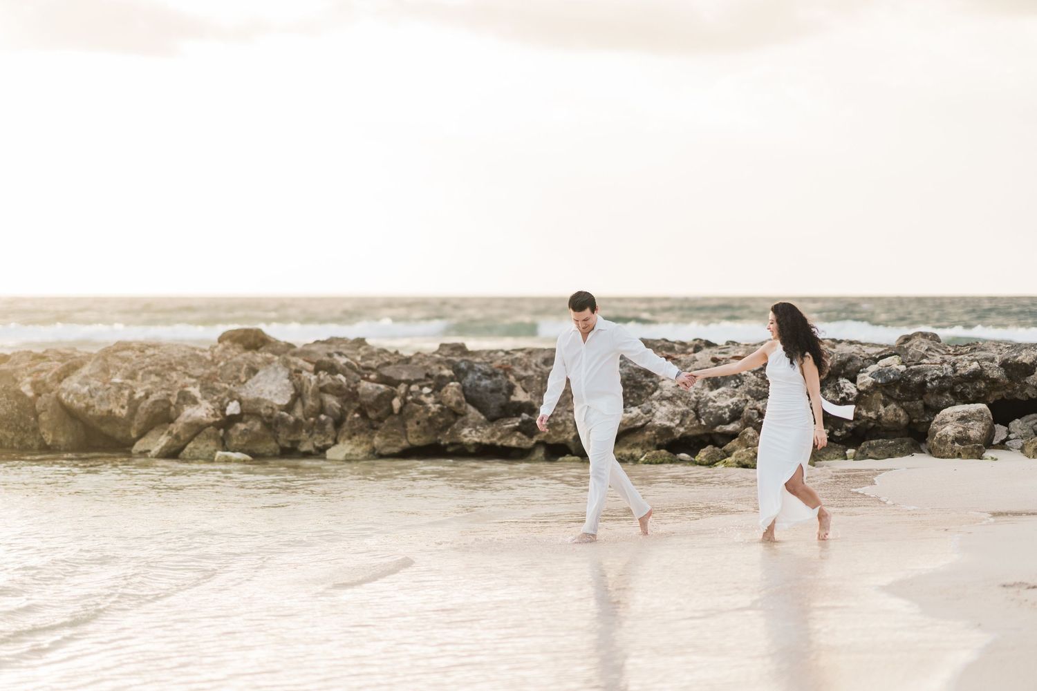 Couple in white outfits holding hands and walking barefoot on a sandy beach with rocky shoreline at sunset.