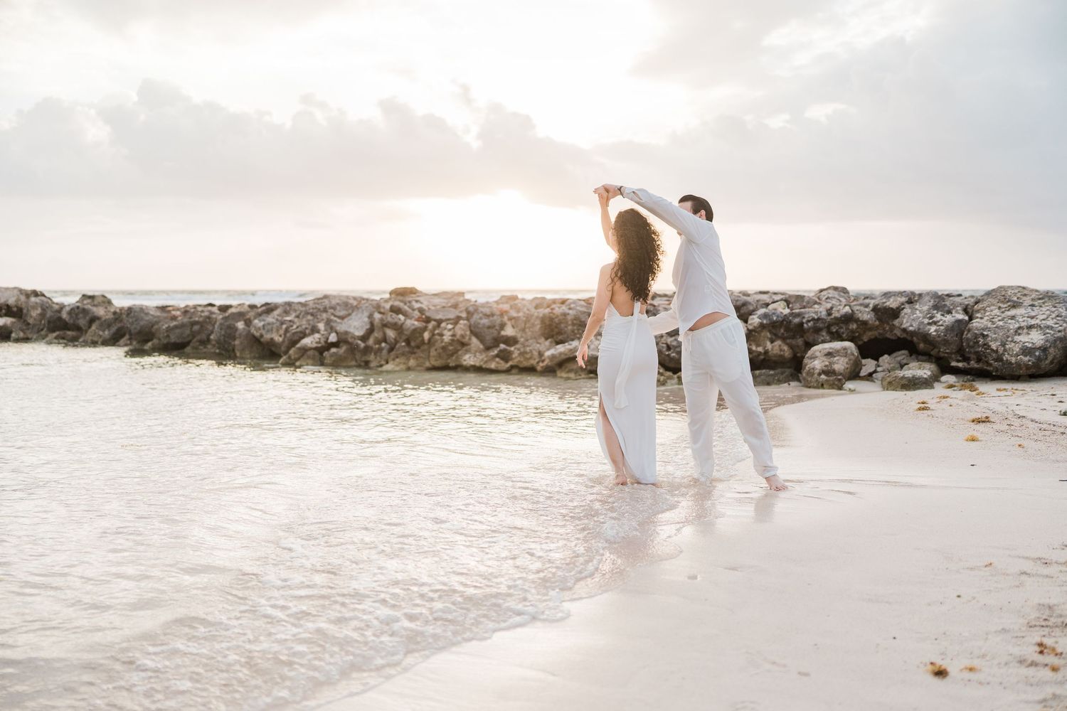 Couple in white outfits dancing on a tropical beach at sunset near rocky shoreline with gentle waves.
