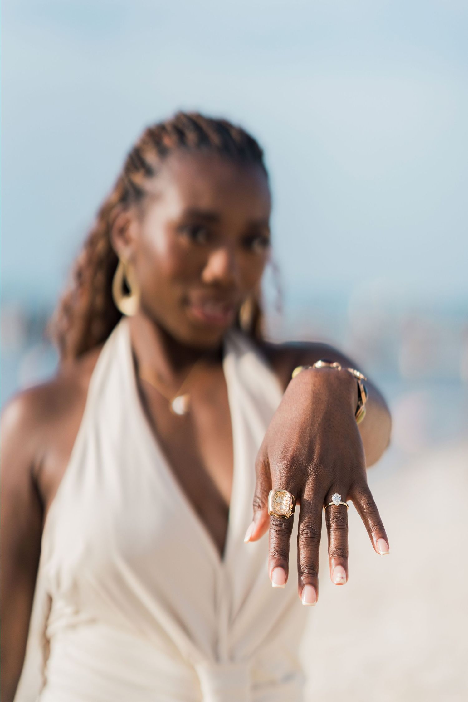 A Black woman in a white halter dress shows off gold rings on her hand at the beach.