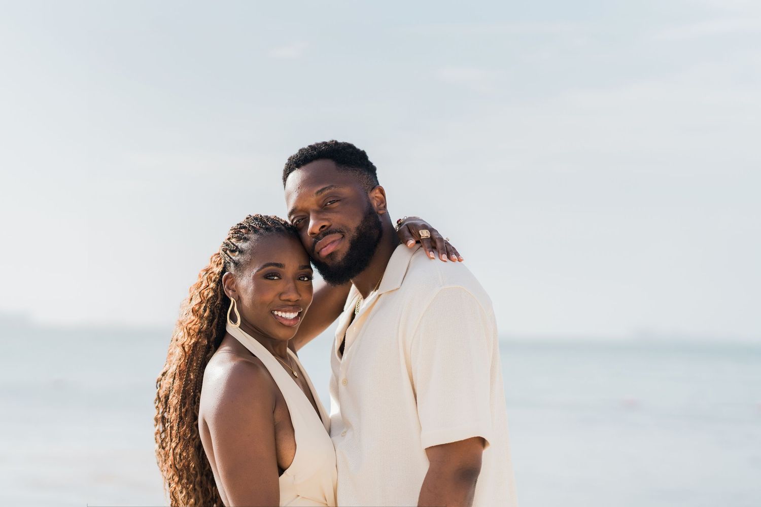 Happy couple embracing on a beach, smiling warmly in matching cream outfits with ocean backdrop.