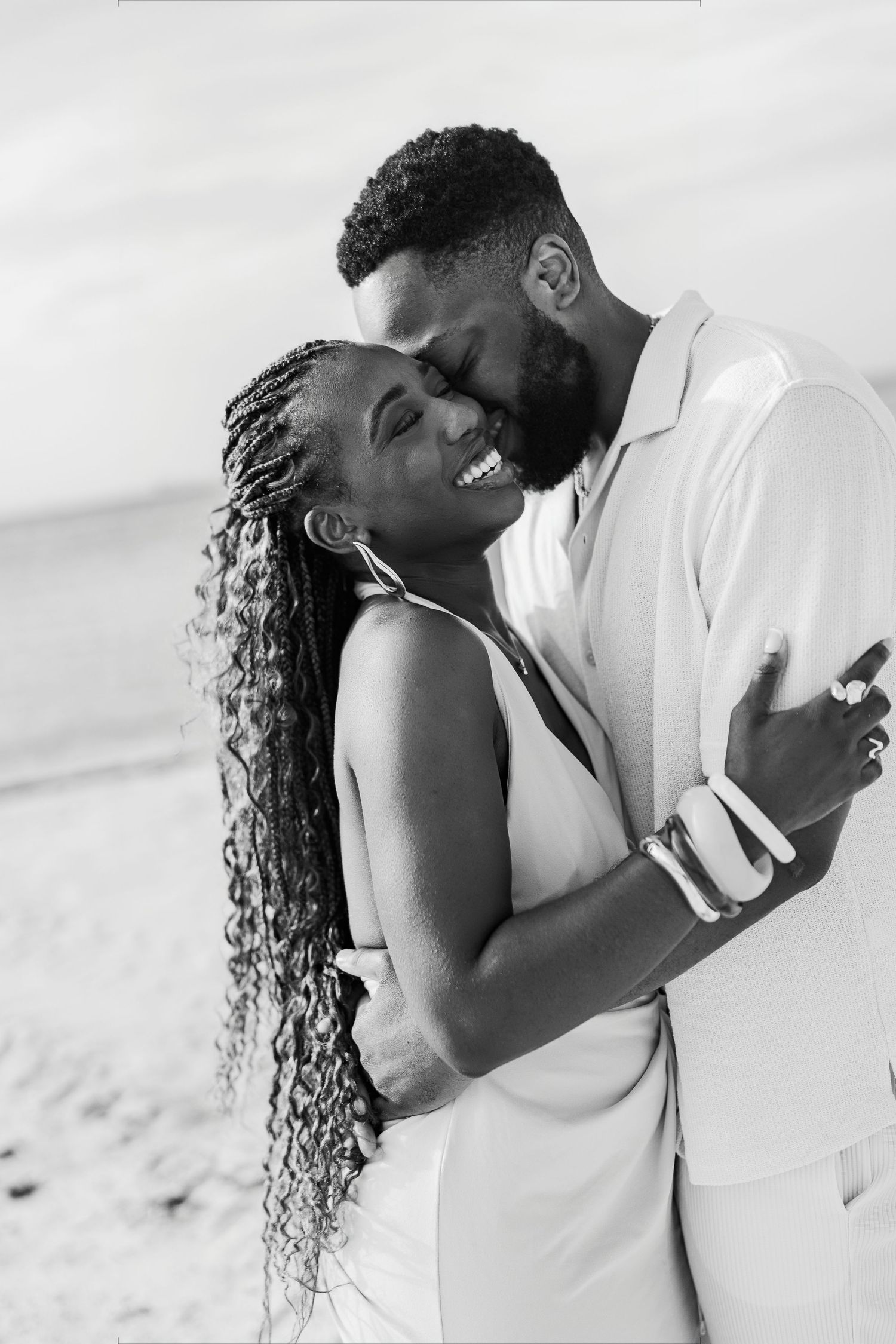 Black and white photo of a joyful couple embracing on a beach, woman smiling with braided hair, man in white suit.