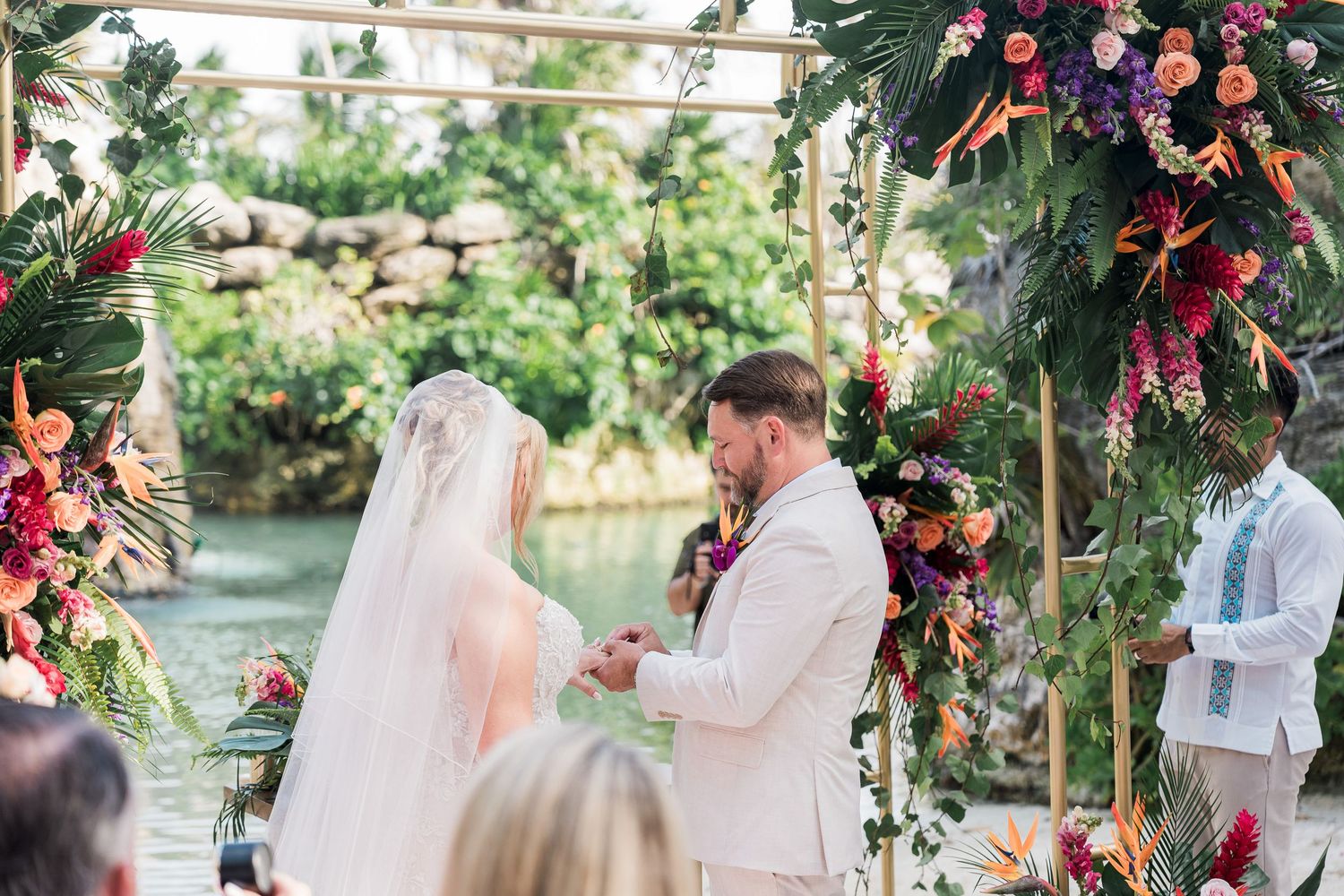 Couple exchanging vows at tropical outdoor wedding ceremony with vibrant floral arch and lush greenery backdrop.