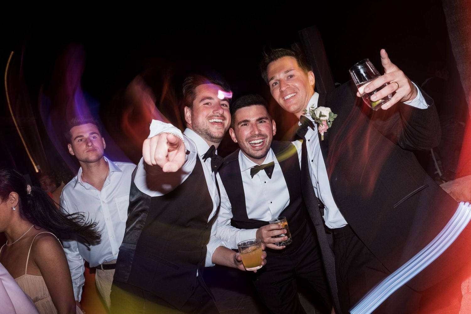 Three men in tuxedos celebrate at a wedding reception, smiling and holding drinks under colorful party lights. TRS Yucatán Wedding