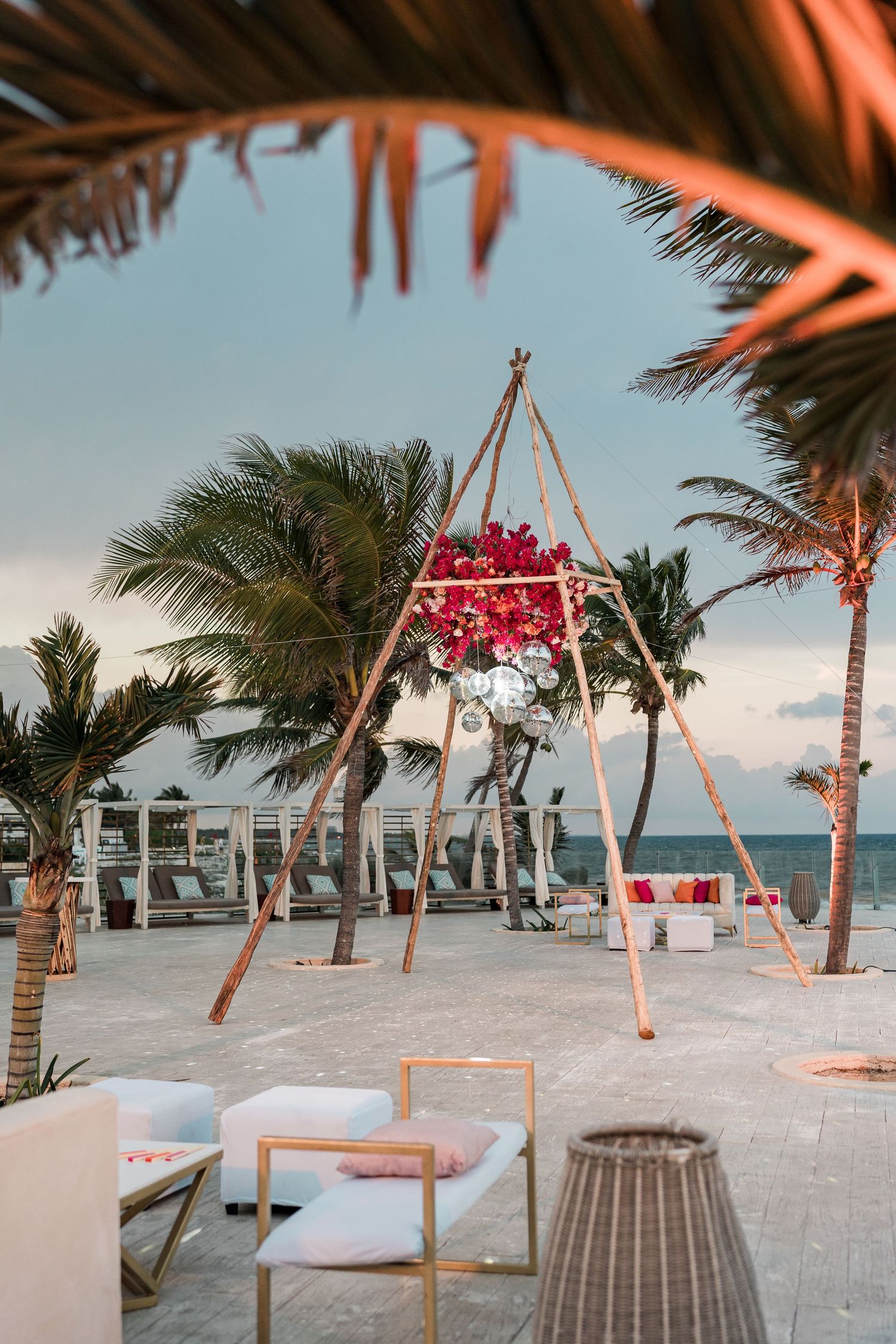 Tropical beach wedding setup with bamboo teepee floral installation, white lounge furniture, and palm trees at sunset.