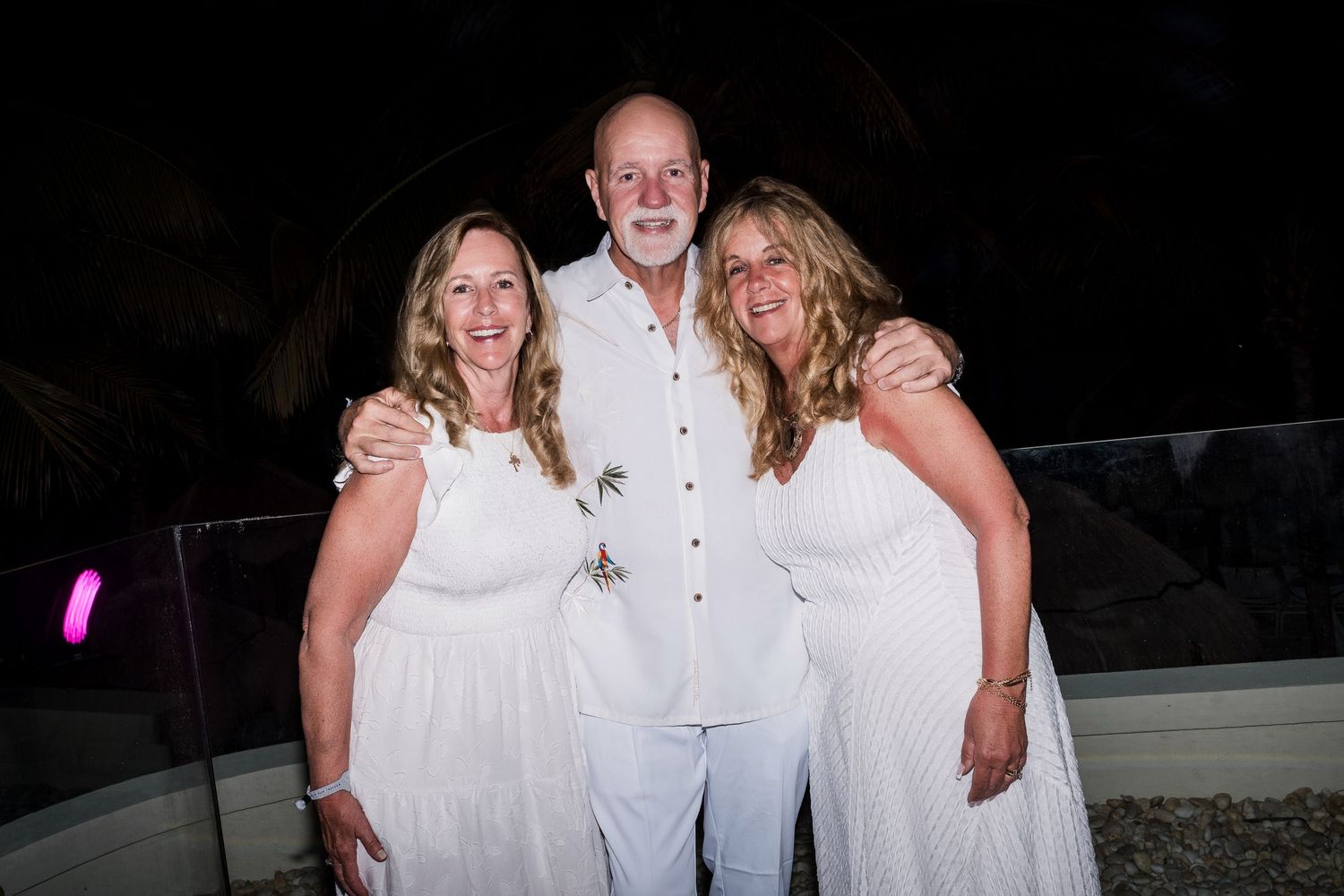 Three people in white outfits smiling together at an outdoor evening event.
