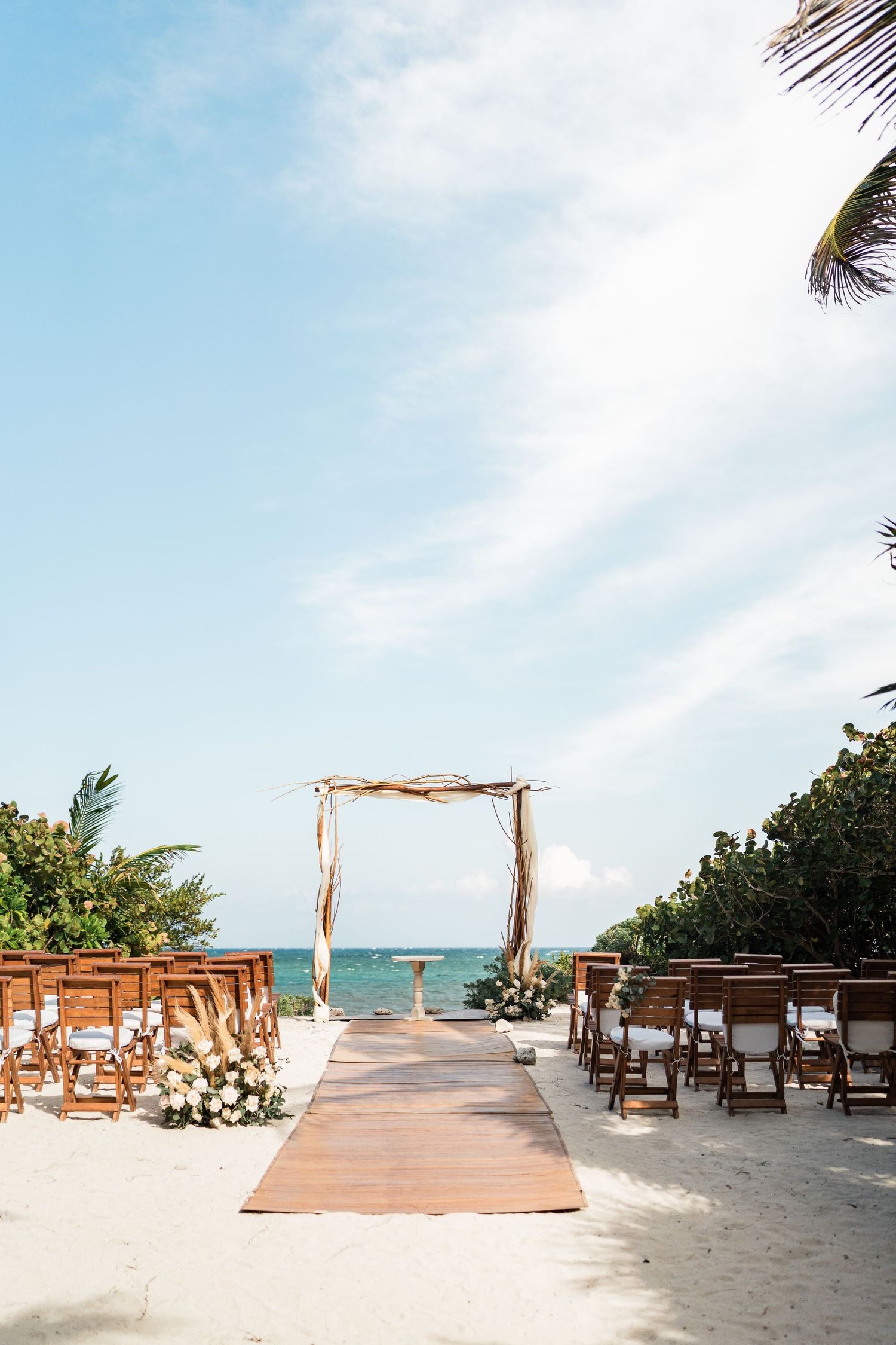 Beach wedding ceremony setup with wooden arch, floral arrangements, and chairs on white sand overlooking the ocean at Akumal Bay Resort