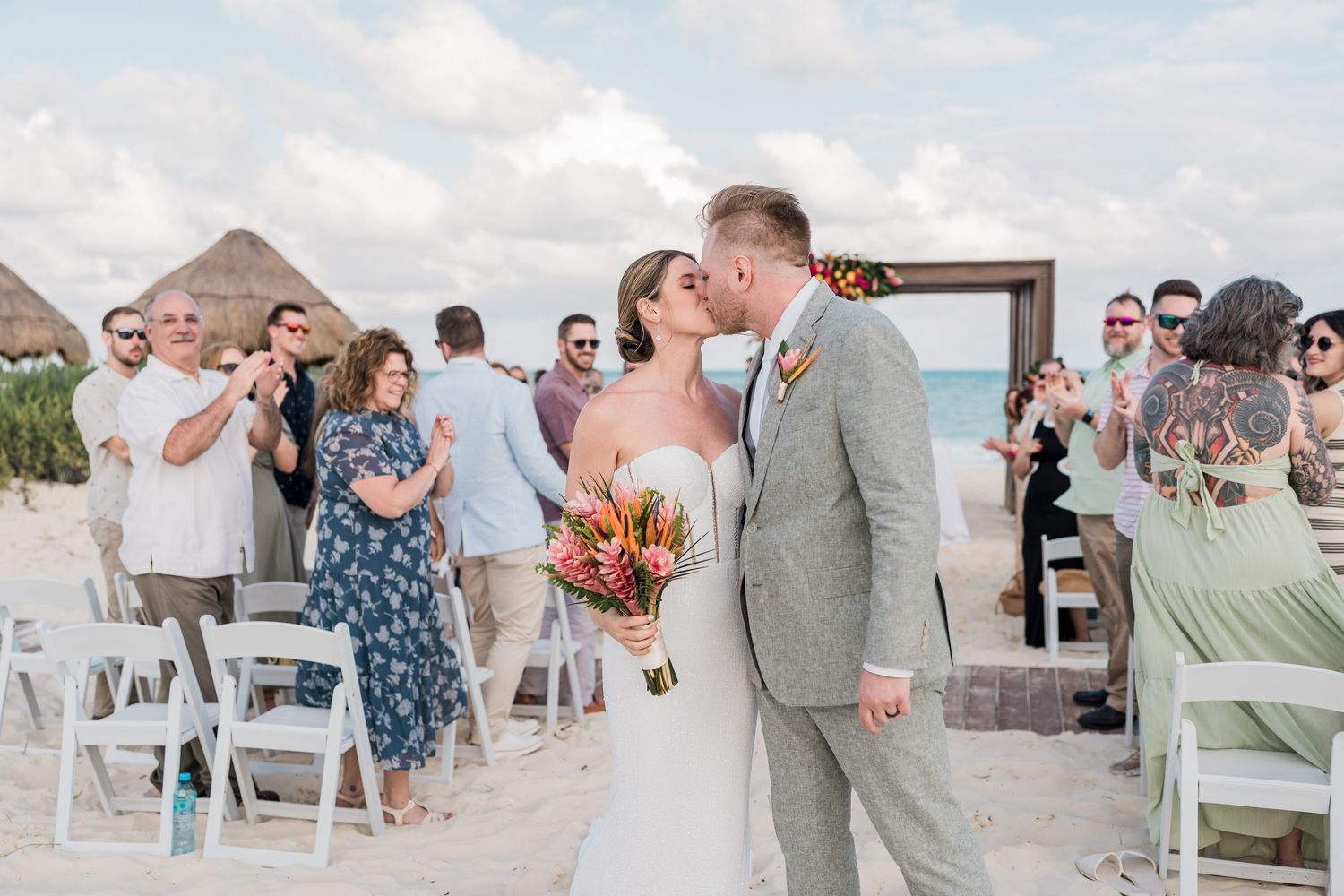 Bride and groom share a kiss on a sandy beach wedding ceremony with guests celebrating in the background.