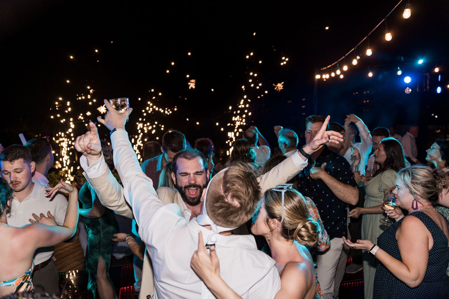 Guests celebrate at a lively outdoor wedding reception with sparklers and string lights at night.