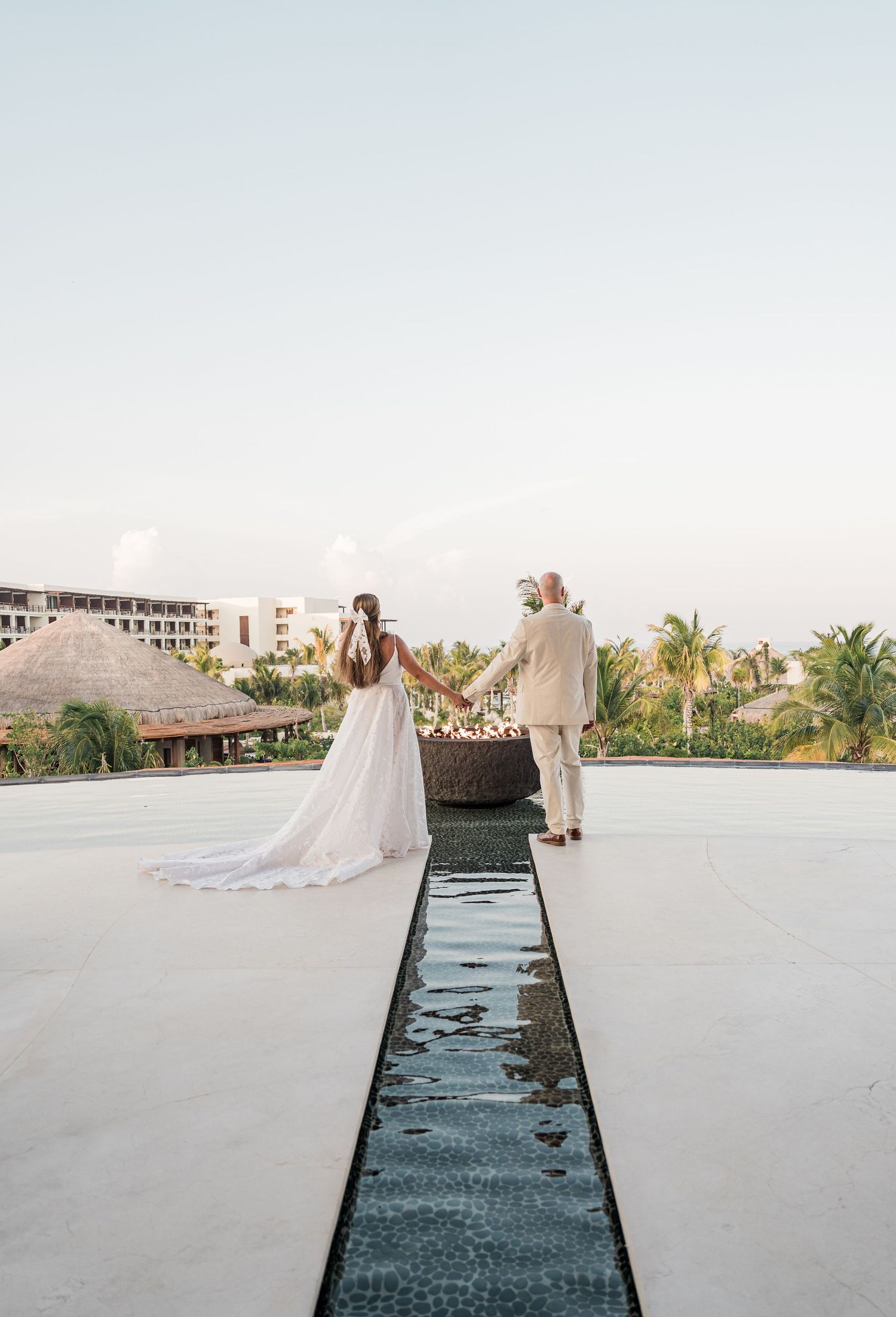Bride and groom hold hands on a rooftop terrace at a tropical resort, surrounded by palm trees and thatched buildings.