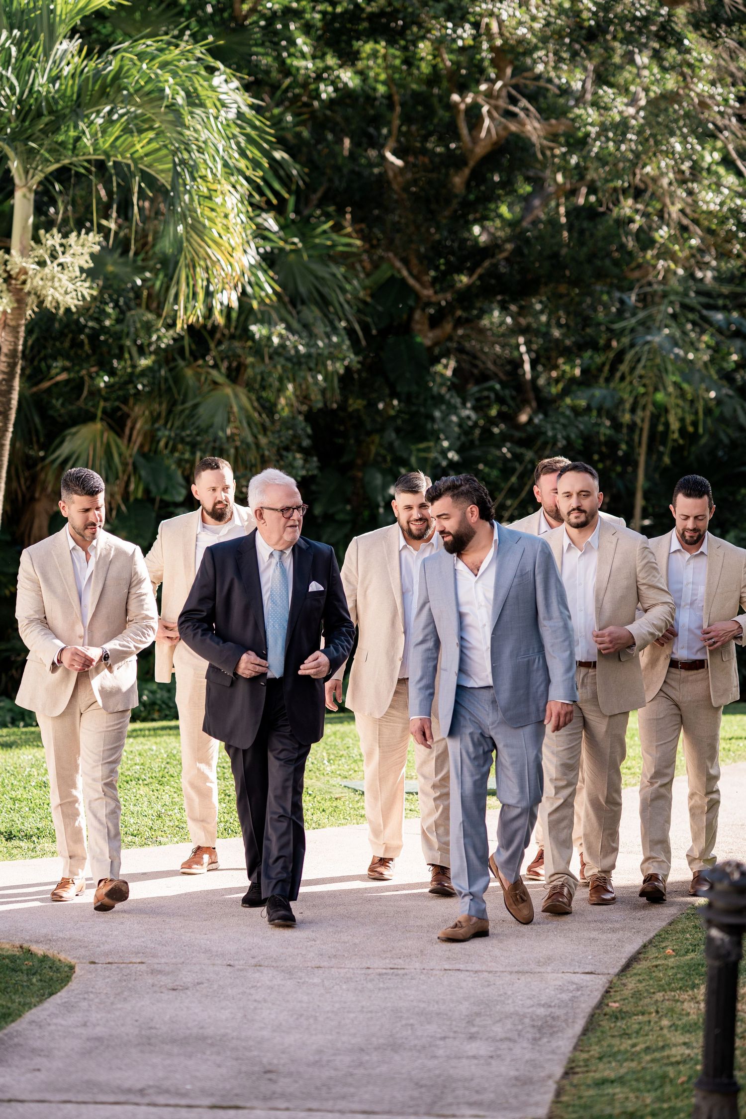 Groom and groomsmen in tan suits walk outdoors on a sunny day surrounded by lush tropical greenery.