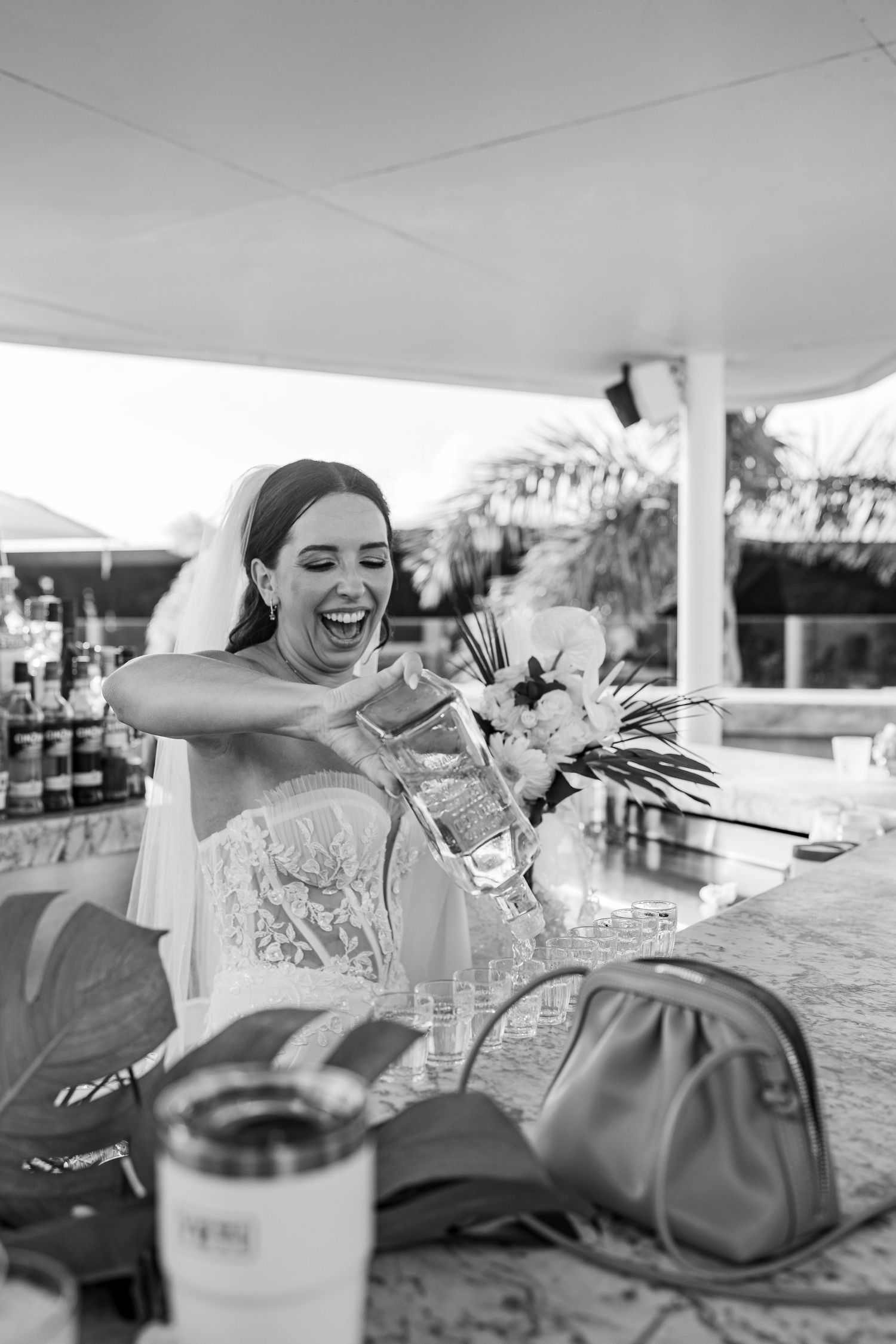 Black and white photo of a laughing bride pouring a drink at an outdoor bar, holding a floral bouquet on her wedding day.