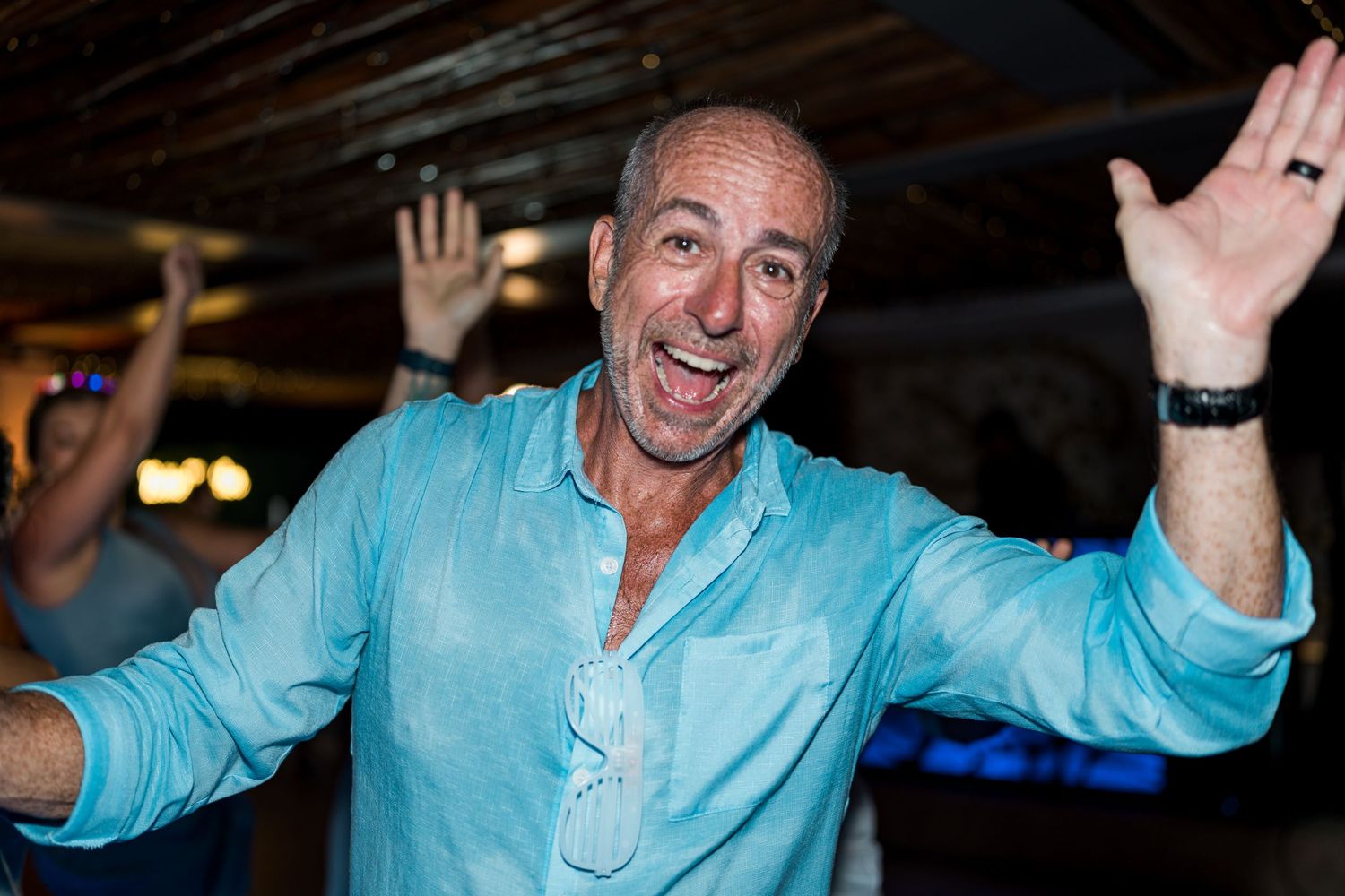 Enthusiastic man in turquoise shirt celebrating with arms raised on the wedding dance floor.