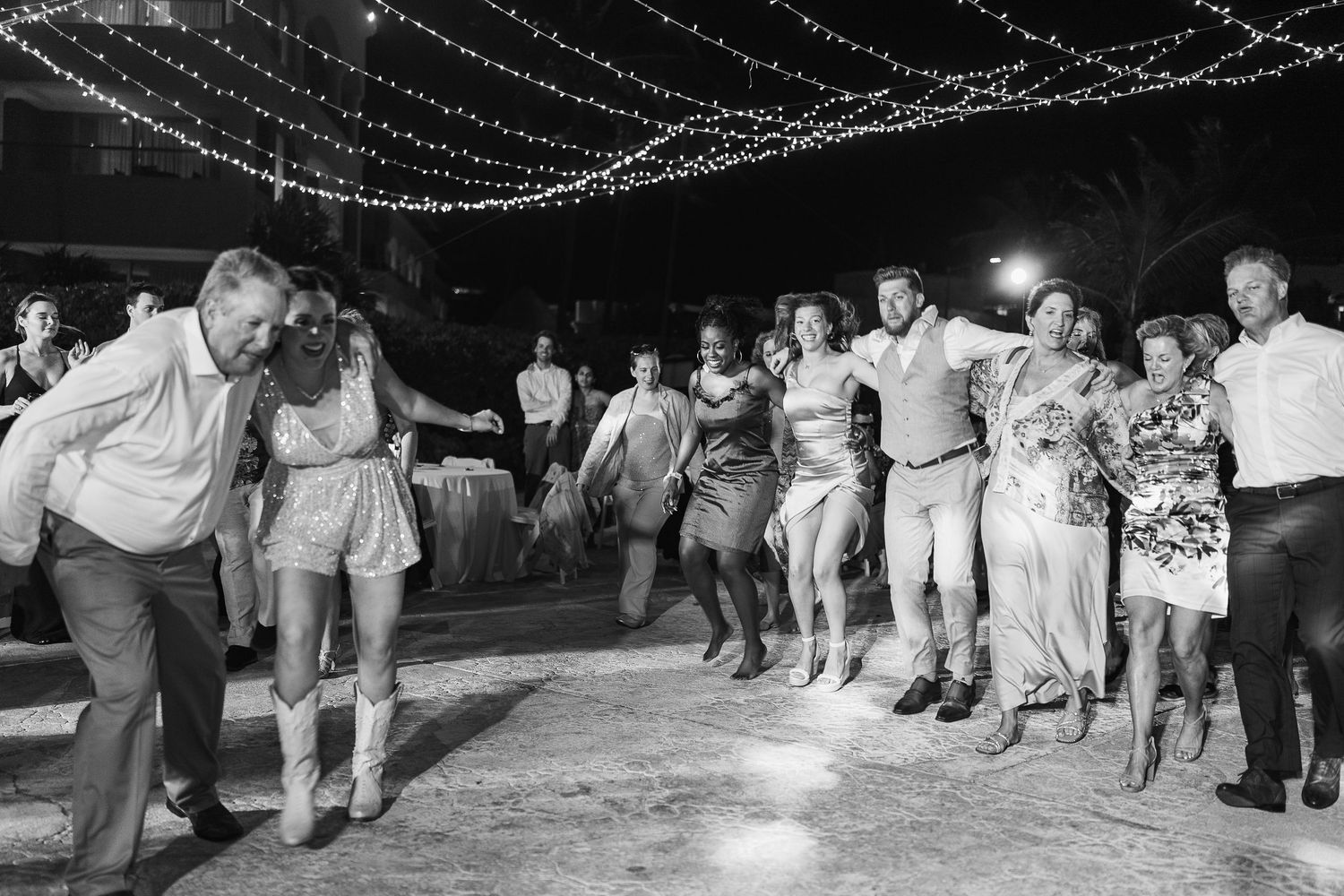 Wedding guests joyfully dance in a line under string lights at an outdoor nighttime reception.