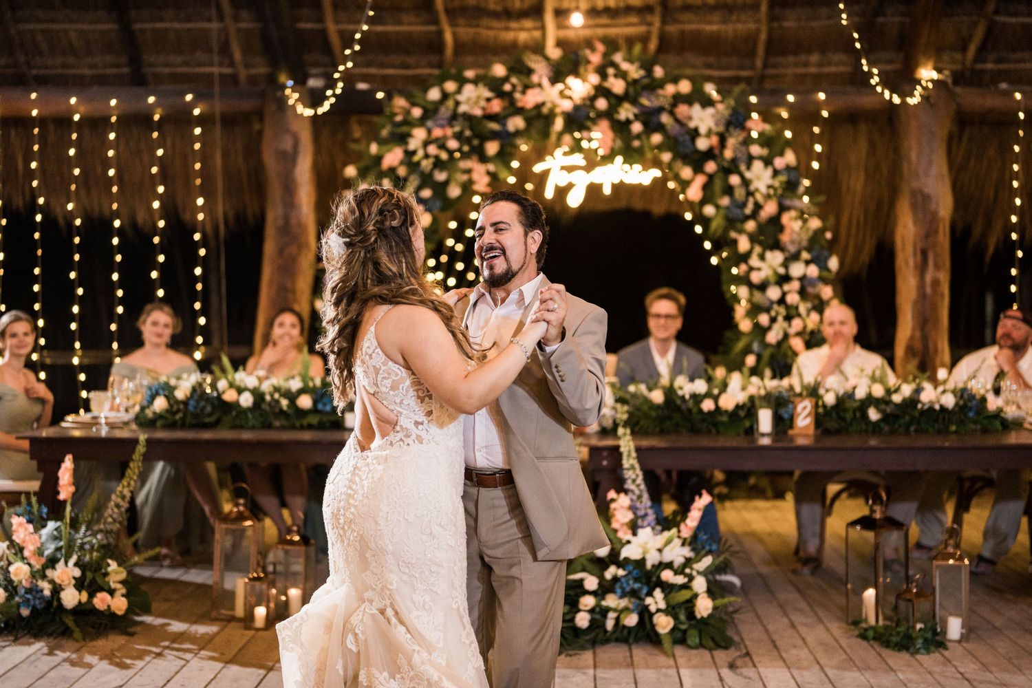 Bride and groom share first dance at rustic wedding reception with floral arch and string lights.