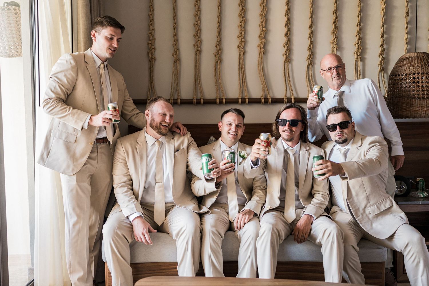 Groom and groomsmen in matching beige suits relaxing on a sofa holding drinks before the wedding.