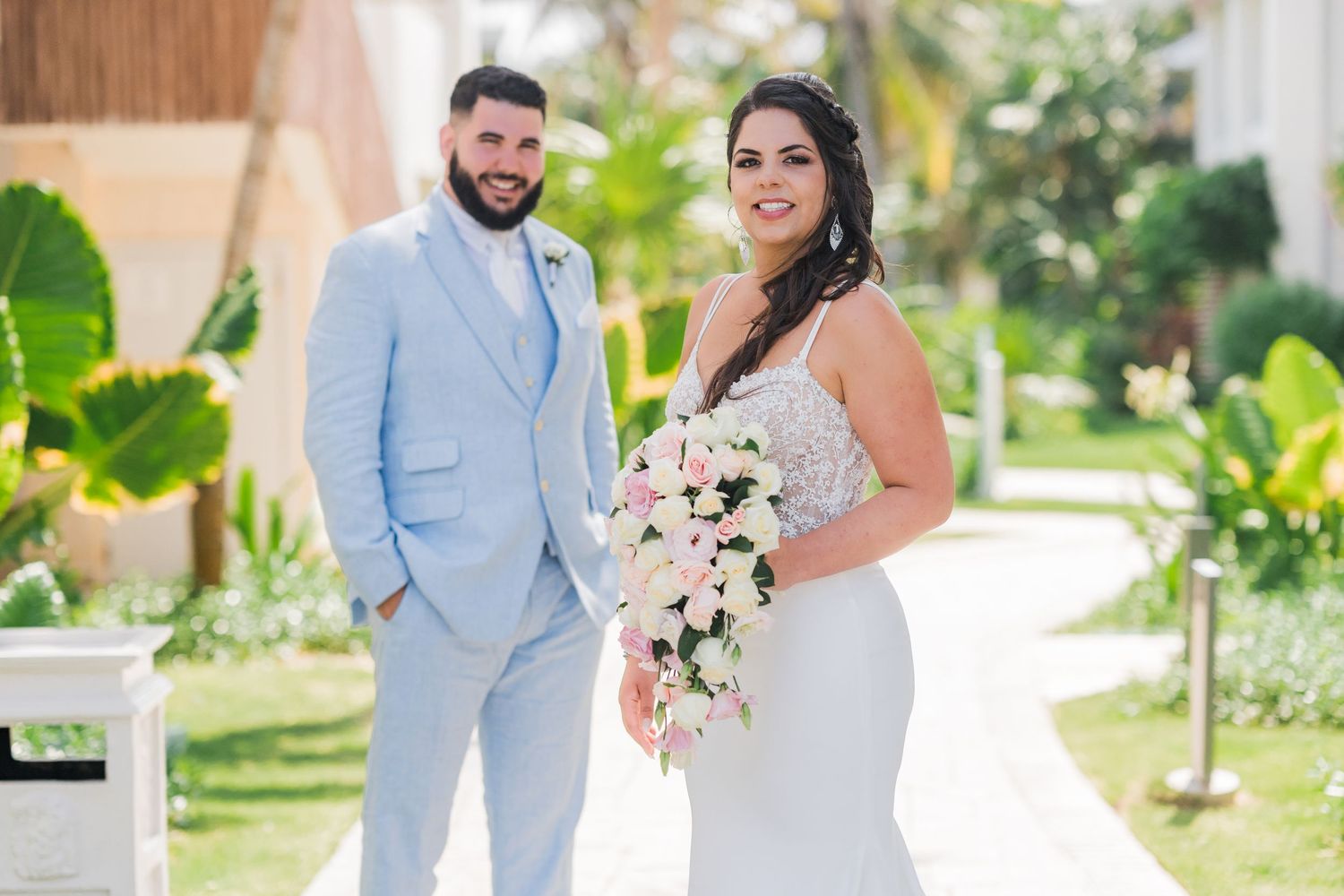 Smiling bride and groom pose together at tropical resort with lush greenery and palm trees.