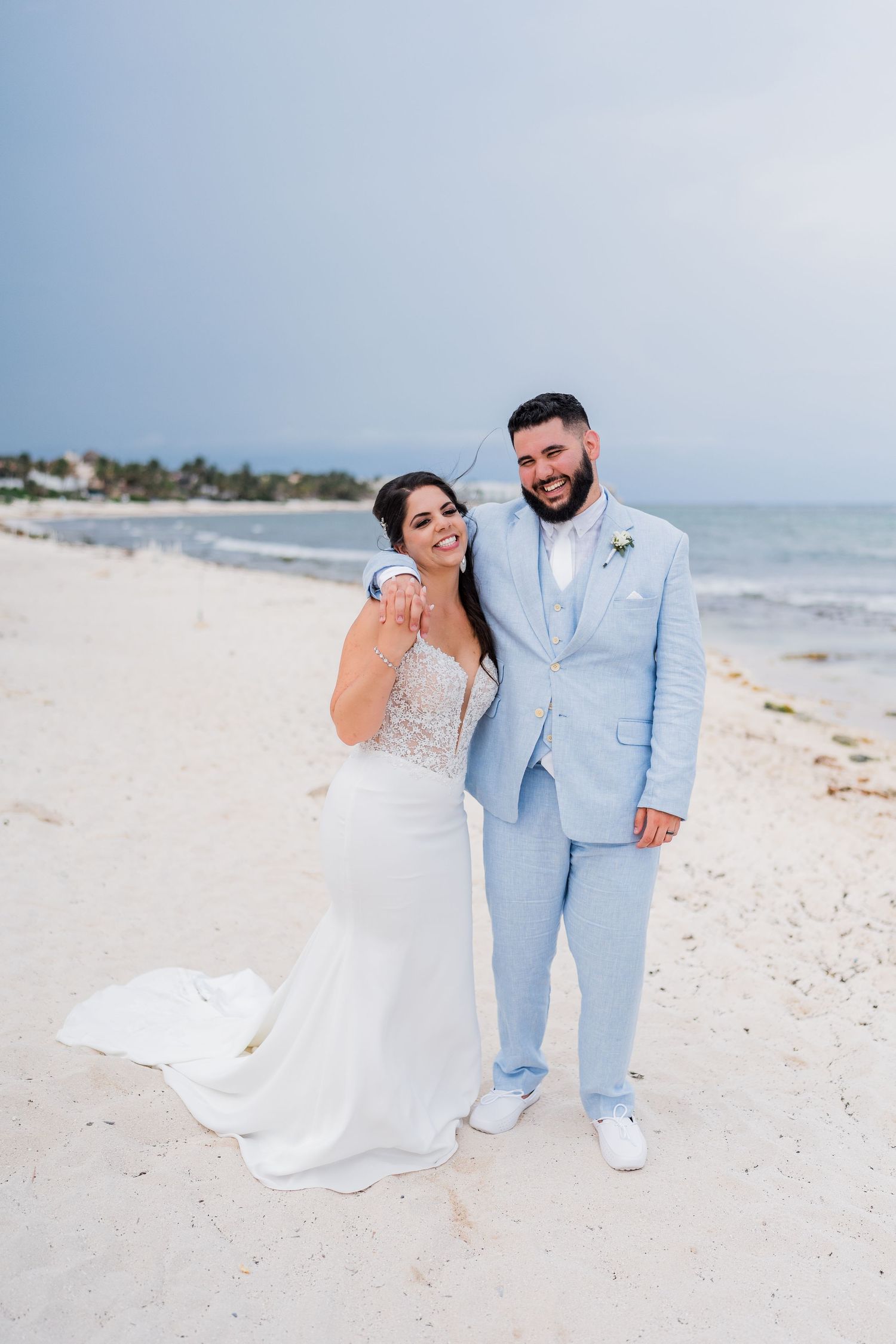 Bride and groom posing happily on a white sand beach, groom in light blue suit, bride in elegant white wedding gown.