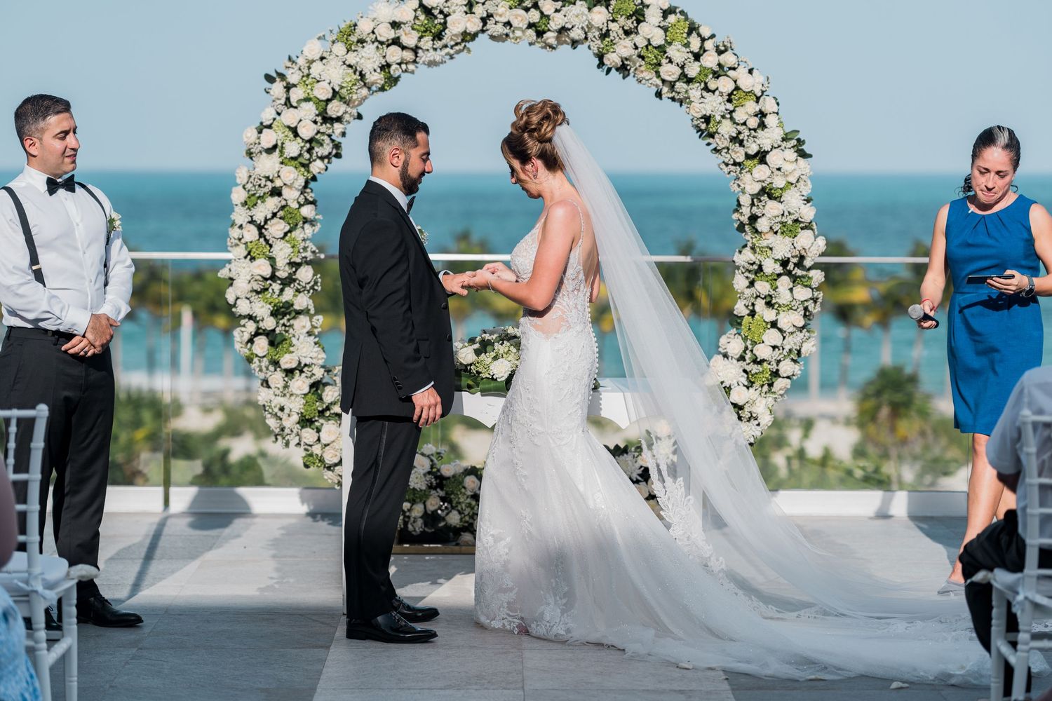 Bride and groom hold hands during vows under white rose arch at seaside wedding.