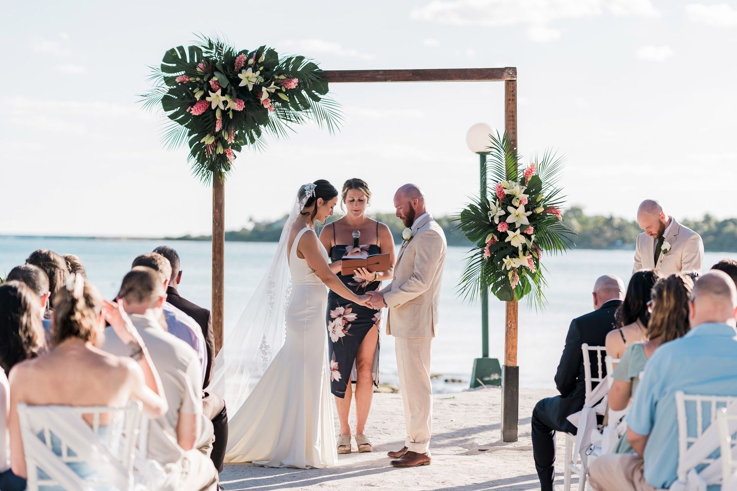 Couple exchanging vows at a tropical outdoor wedding ceremony on a waterfront deck under a wooden arch with floral decor.