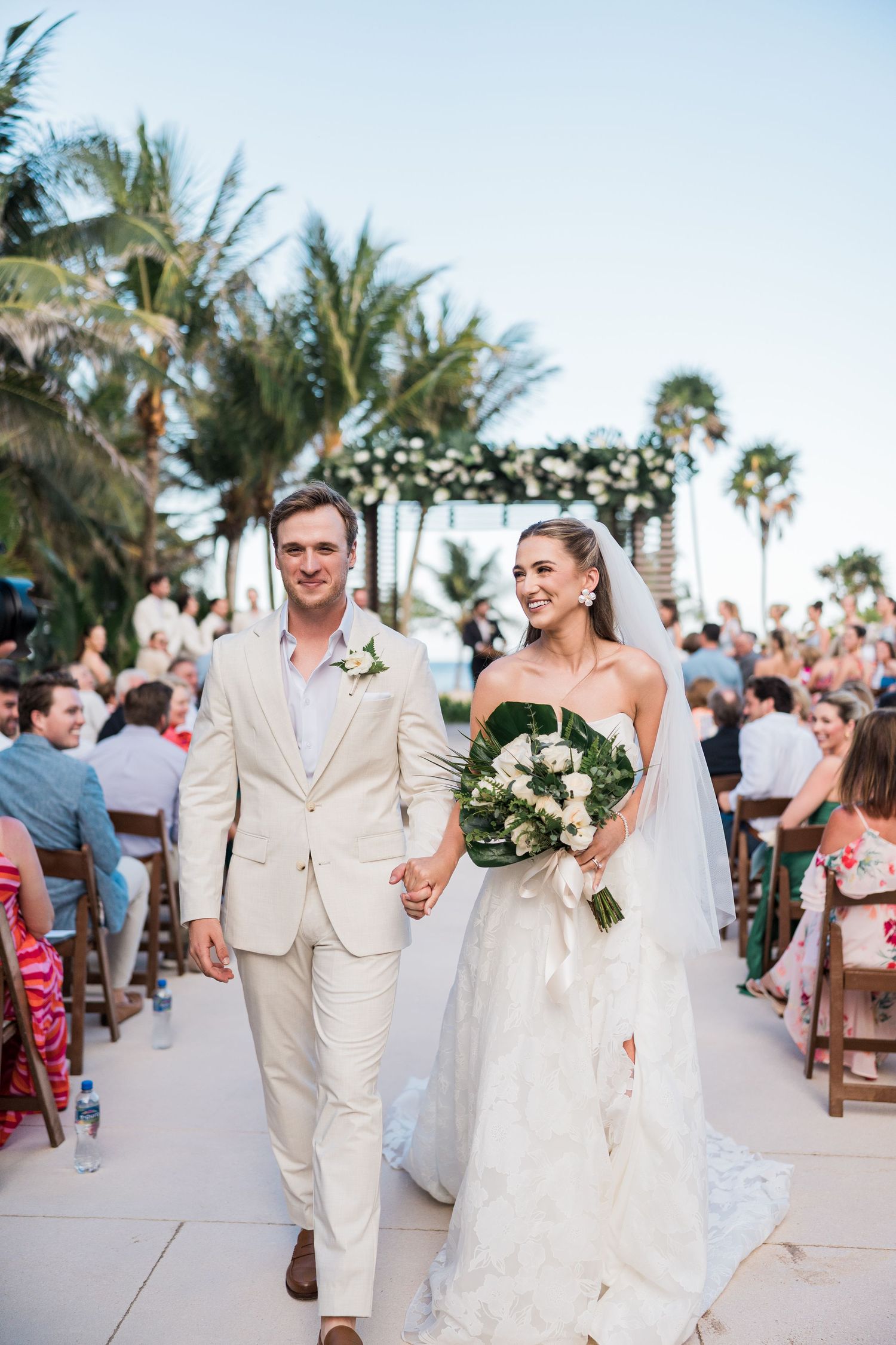 Bride and groom walk down the aisle at an outdoor beach wedding ceremony surrounded by palm trees and guests.