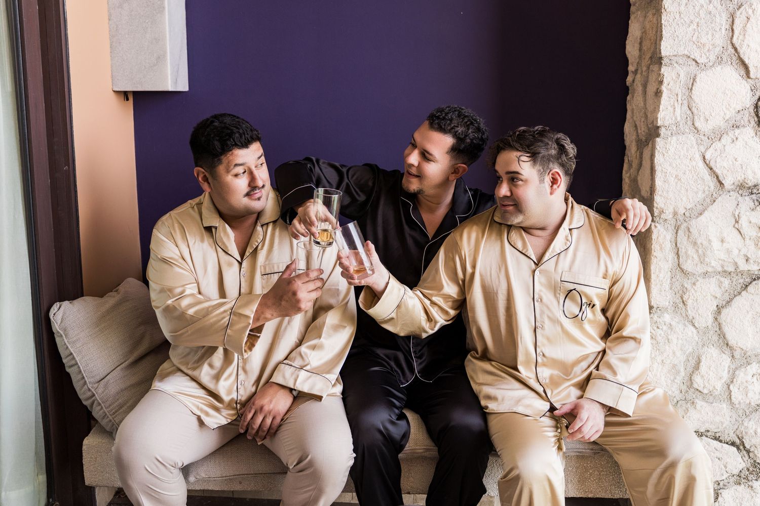 Xcaret Arte Wedding - Three men in satin pajamas laughing and toasting drinks together on a couch in a stylish room.