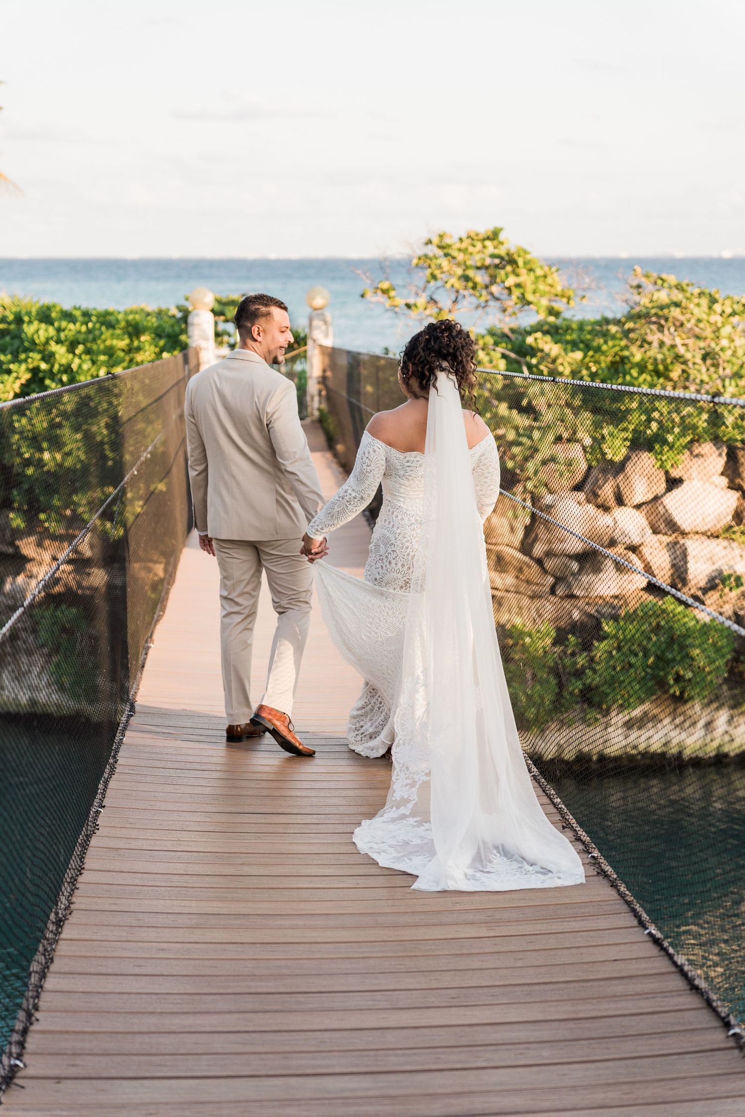 Xcaret Arte Wedding - Bride and groom holding hands on a wooden suspension bridge overlooking the ocean on their wedding day.