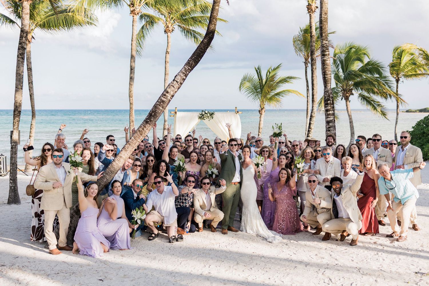 Large wedding group photo on a tropical beach with palm trees, ocean backdrop, bride and groom surrounded by guests.