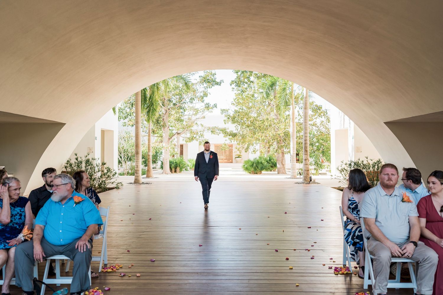 Secrets Akumal Wedding - Groom walks down outdoor wedding aisle under arched venue with seated guests on both sides.