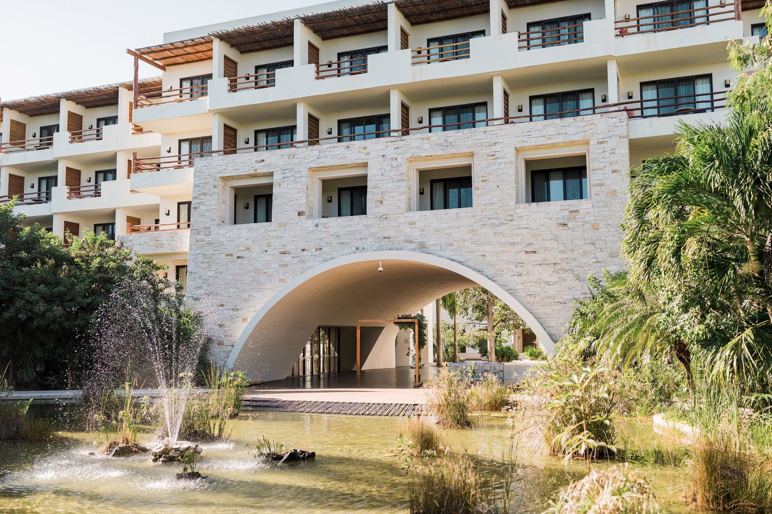 Secrets Akumal Wedding - Modern hotel exterior with stone facade, arched entrance, fountain, and lush gardens under clear sky.