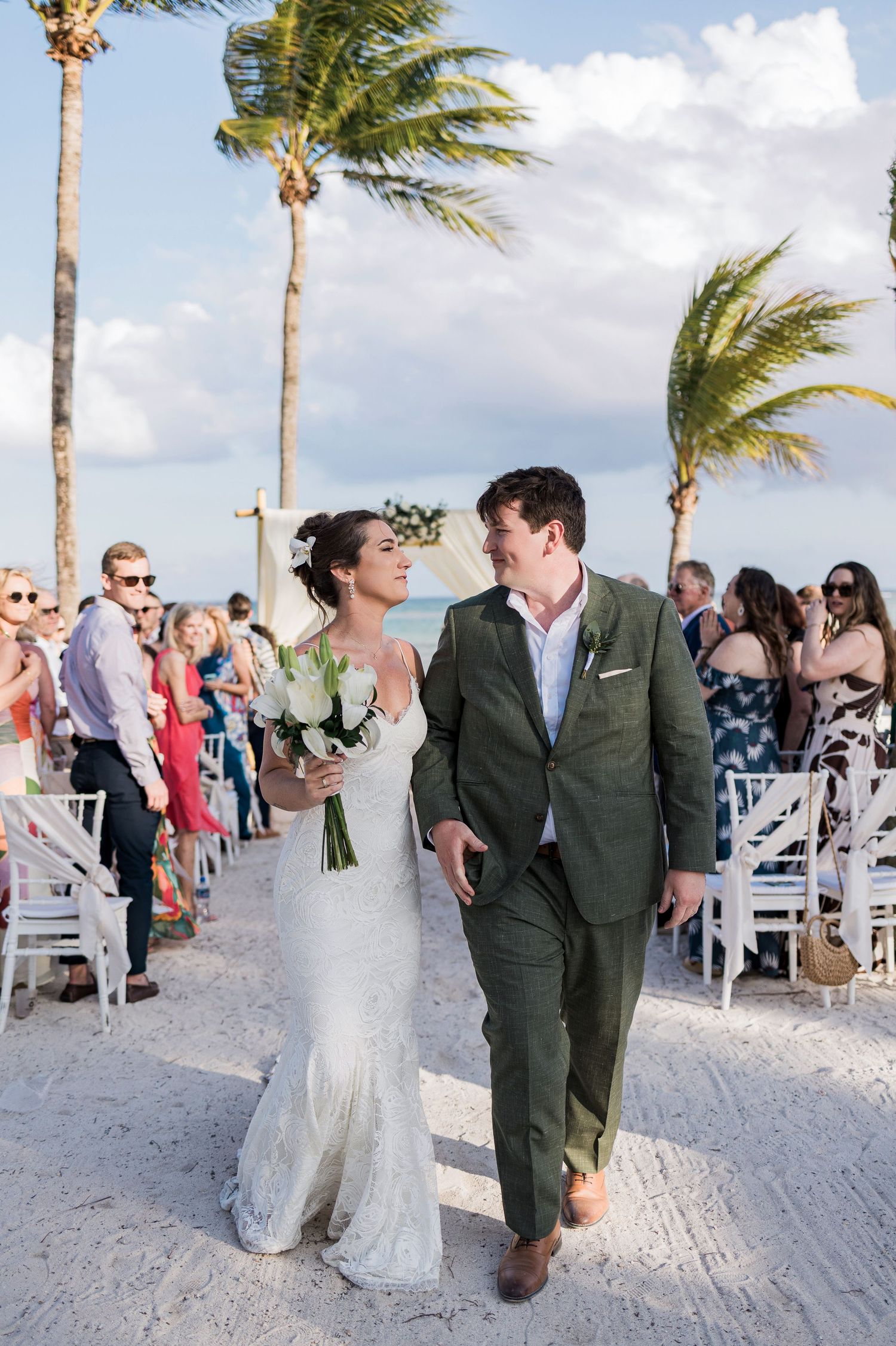 Bride and groom walk down sandy beach aisle after ceremony, palm trees swaying, guests cheering, bride holding white bouquet.