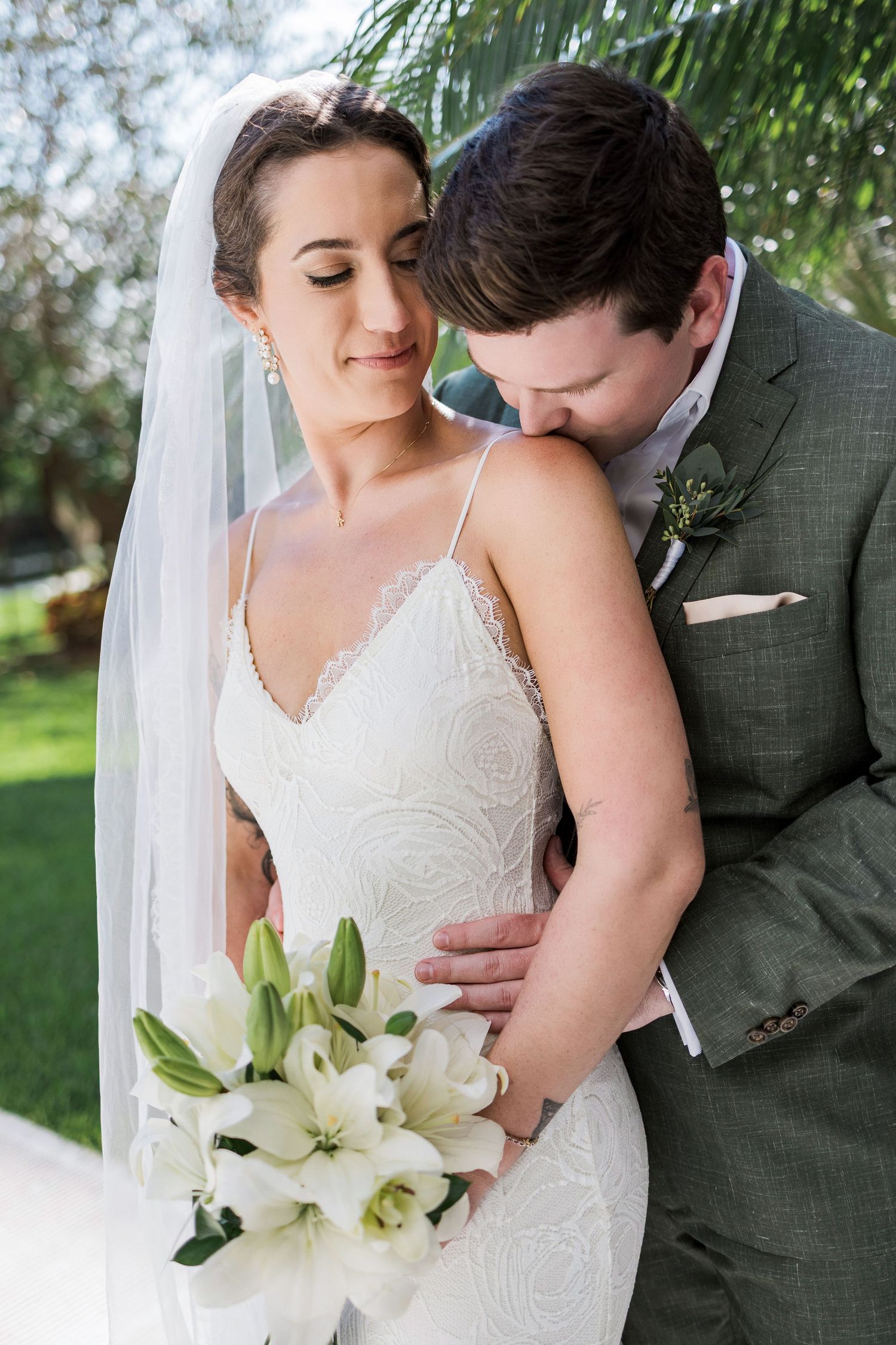Bride in lace gown holding white lily bouquet, groom in sage suit nuzzling her neck during romantic outdoor wedding portrait.