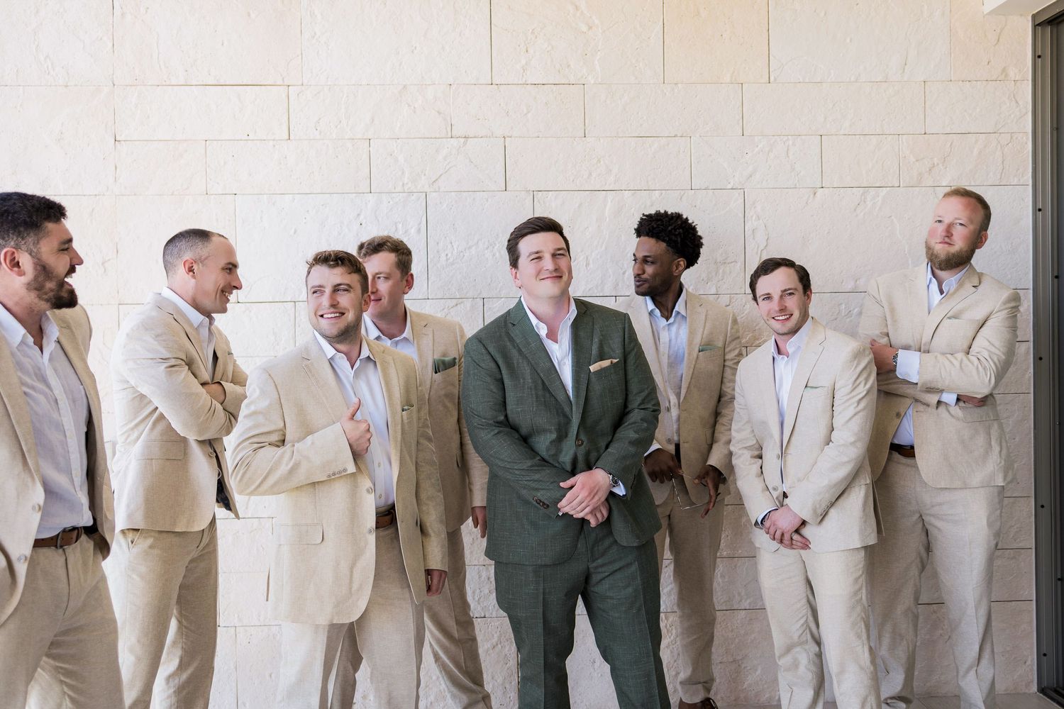 Groom in green suit stands with groomsmen in tan suits laughing together against a stone wall.