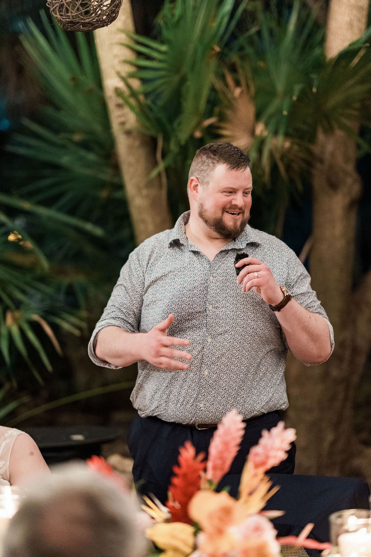 Secrets Akumal Wedding - A bearded man in a patterned shirt gives a speech holding a microphone at a tropical wedding reception.