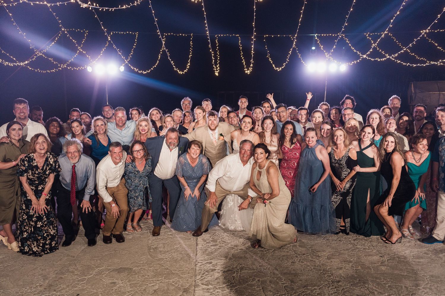 Large wedding party group photo with bride and groom surrounded by guests under string lights at night.
