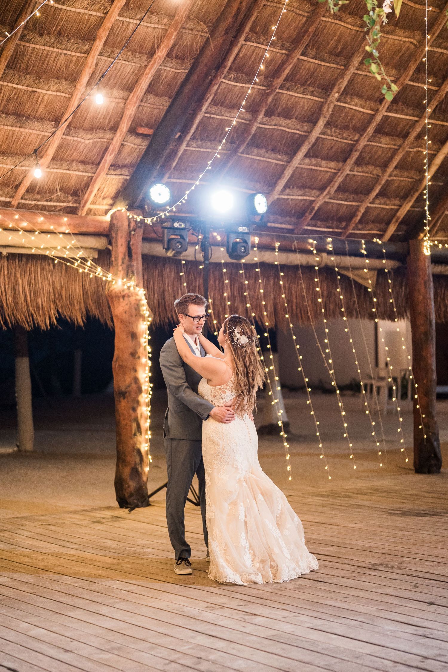 Bride and groom dancing under warm string lights in a rustic thatched-roof palapa wedding reception.