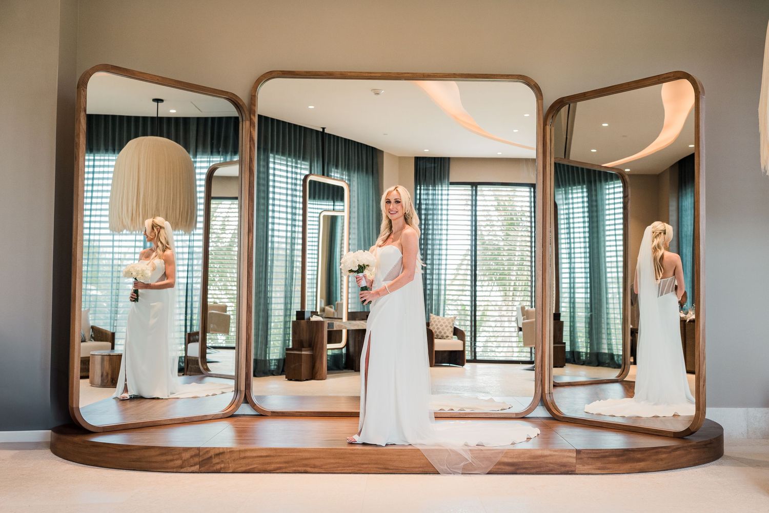 Elegant blonde bride in white gown holding bouquet reflected in a large trifold dressing mirror.