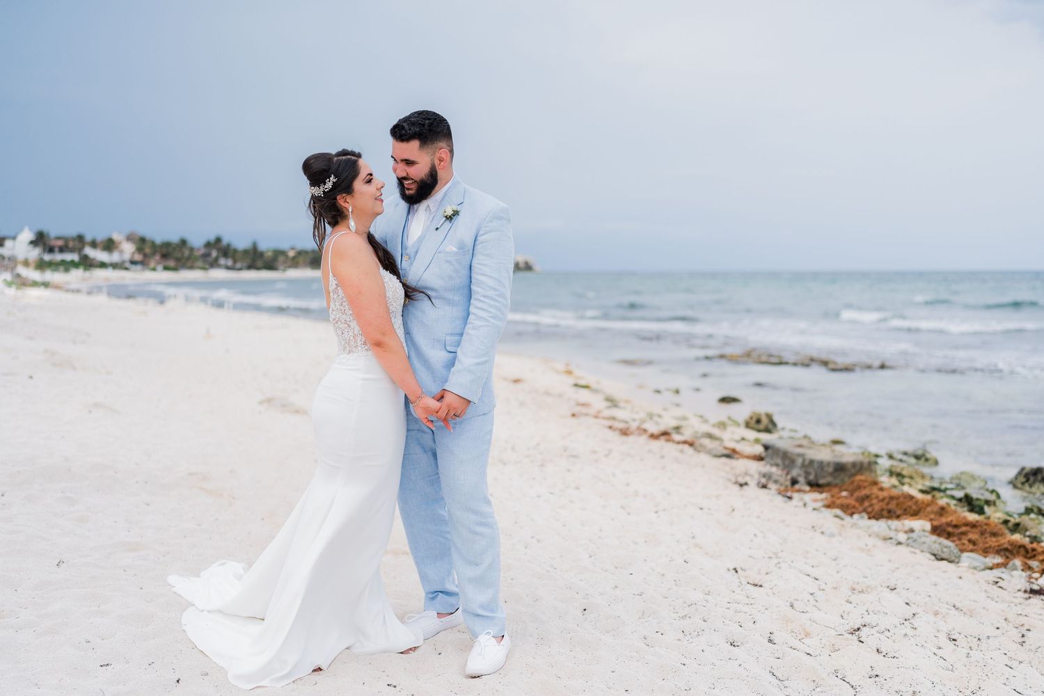 Bride and bearded groom hold hands facing each other on a white sandy beach with ocean backdrop.