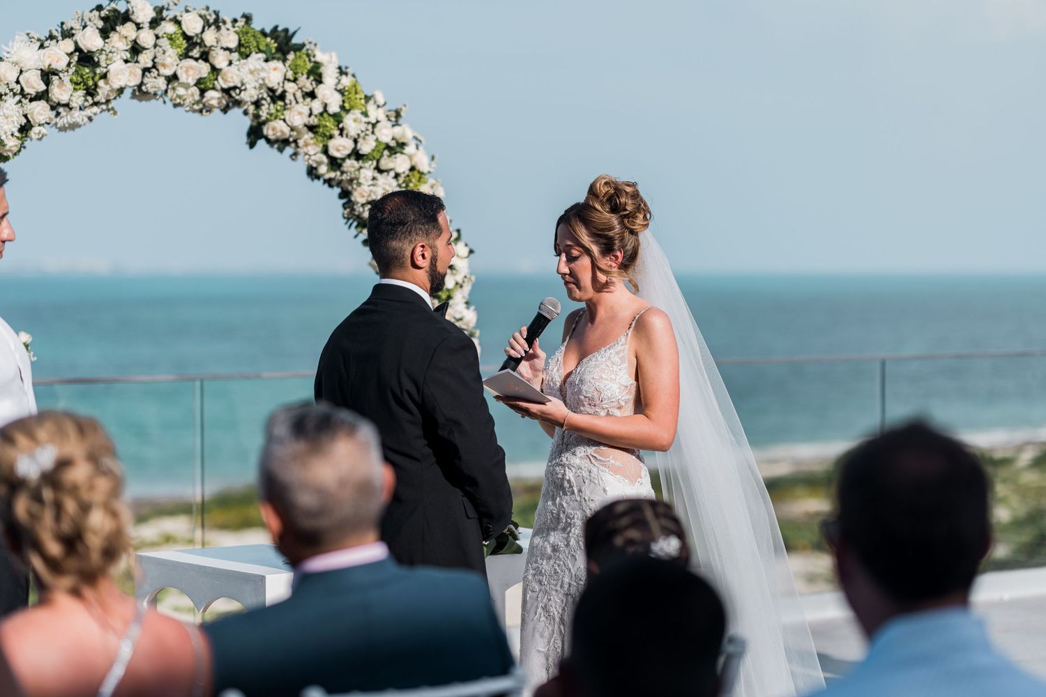 Bride reads wedding vows holding microphone under white rose arch by the sea.