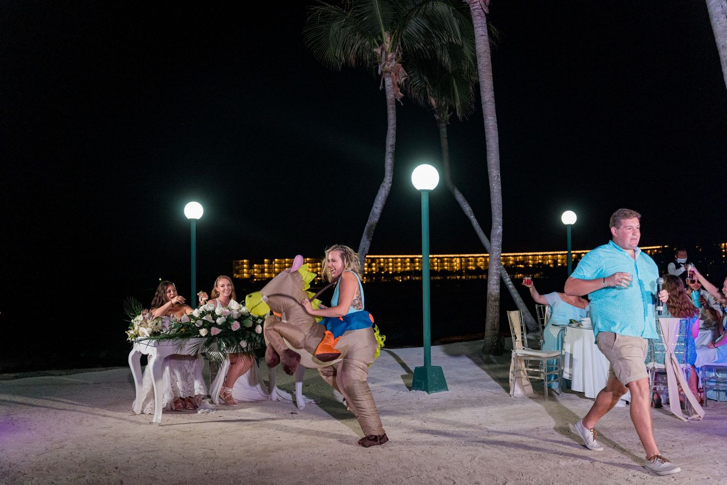 Guests run and dance at an outdoor beach wedding reception at night with palm trees and string lights.