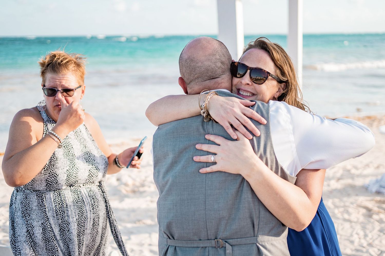 Emotional wedding guests share warm hugs with groom on sandy beach after outdoor ceremony.