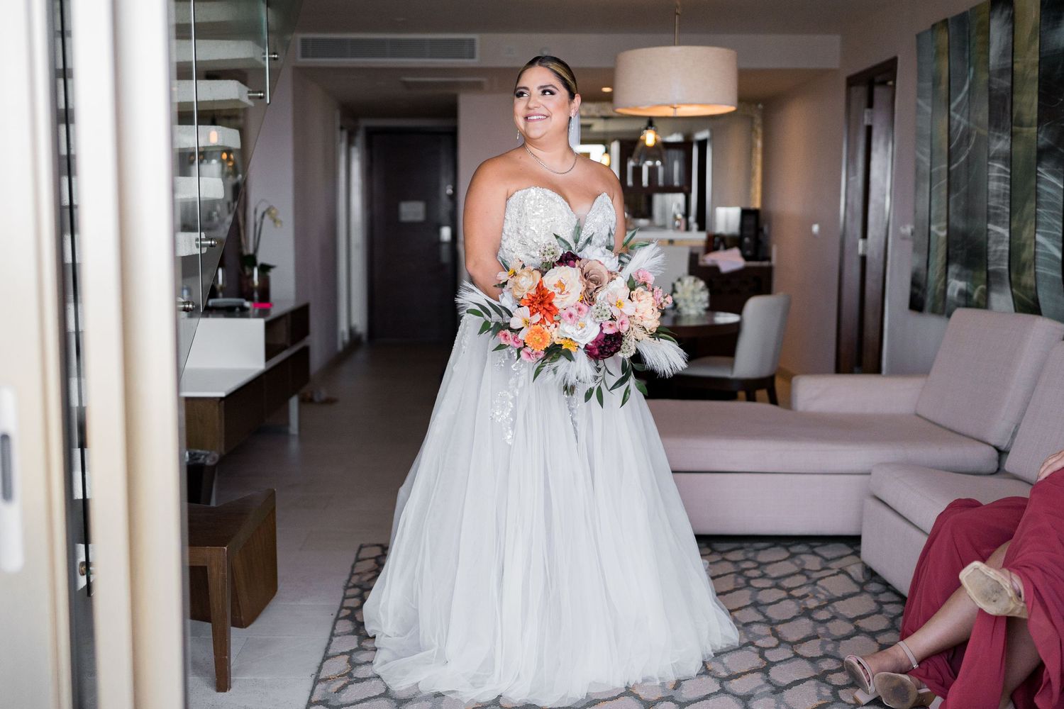 Bride in a strapless tulle ball gown holding a colorful orange and pink bouquet in a hotel suite.