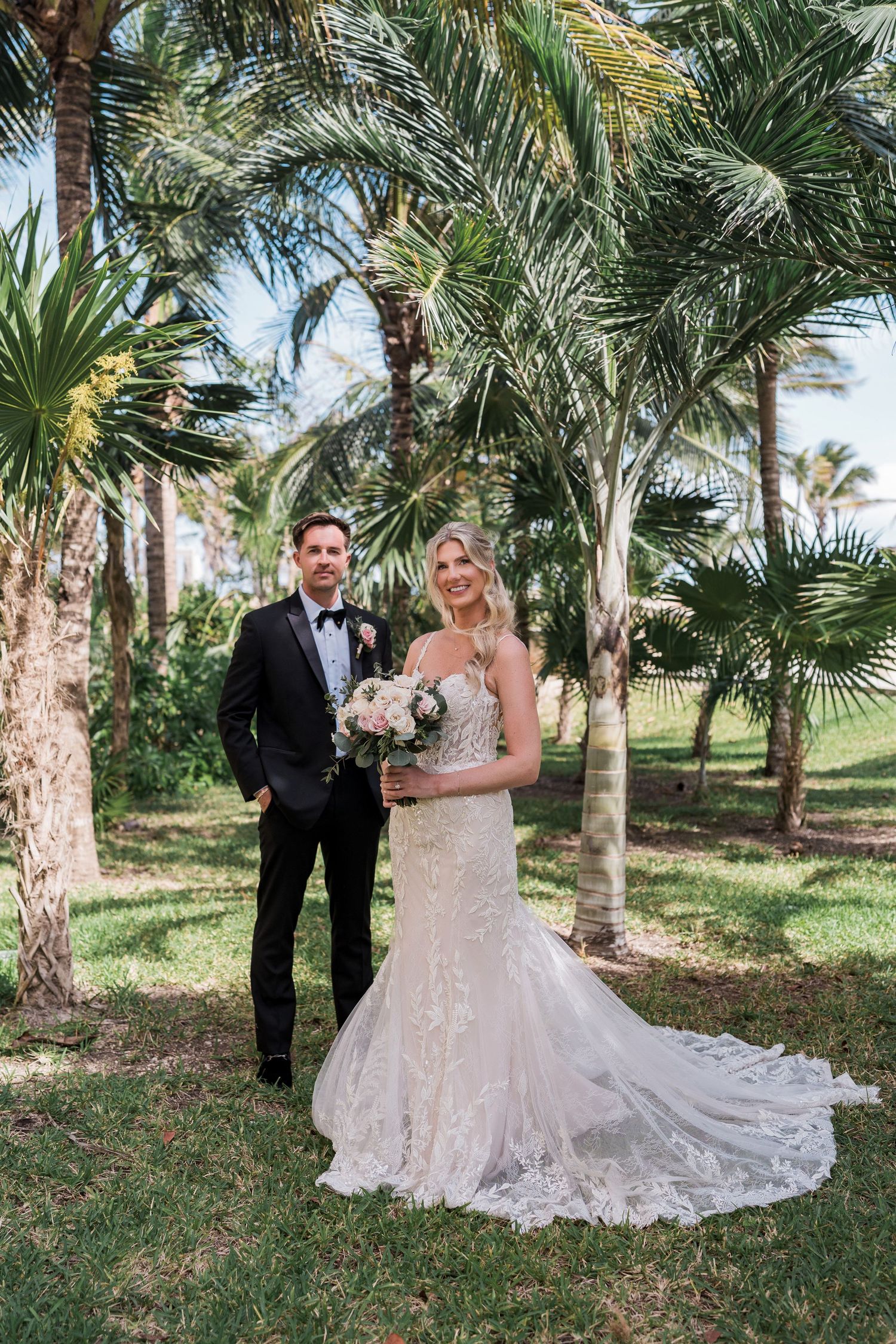Bride in lace mermaid gown holding bouquet stands with tuxedo groom among tropical palm trees on wedding day.