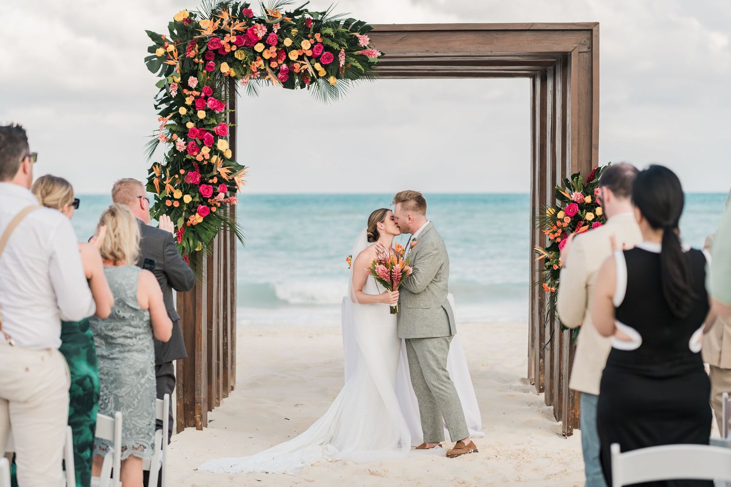Bride and groom share first kiss under floral arch on beach wedding ceremony with ocean backdrop.