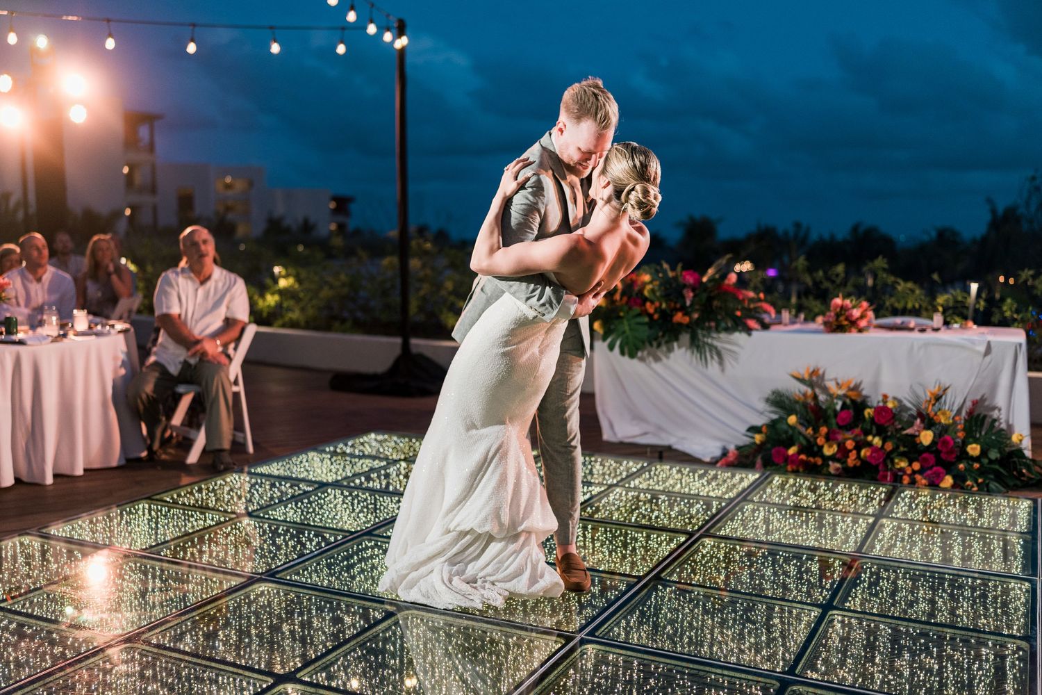 Couple shares romantic first dance on illuminated glass floor at outdoor wedding reception under string lights at dusk.