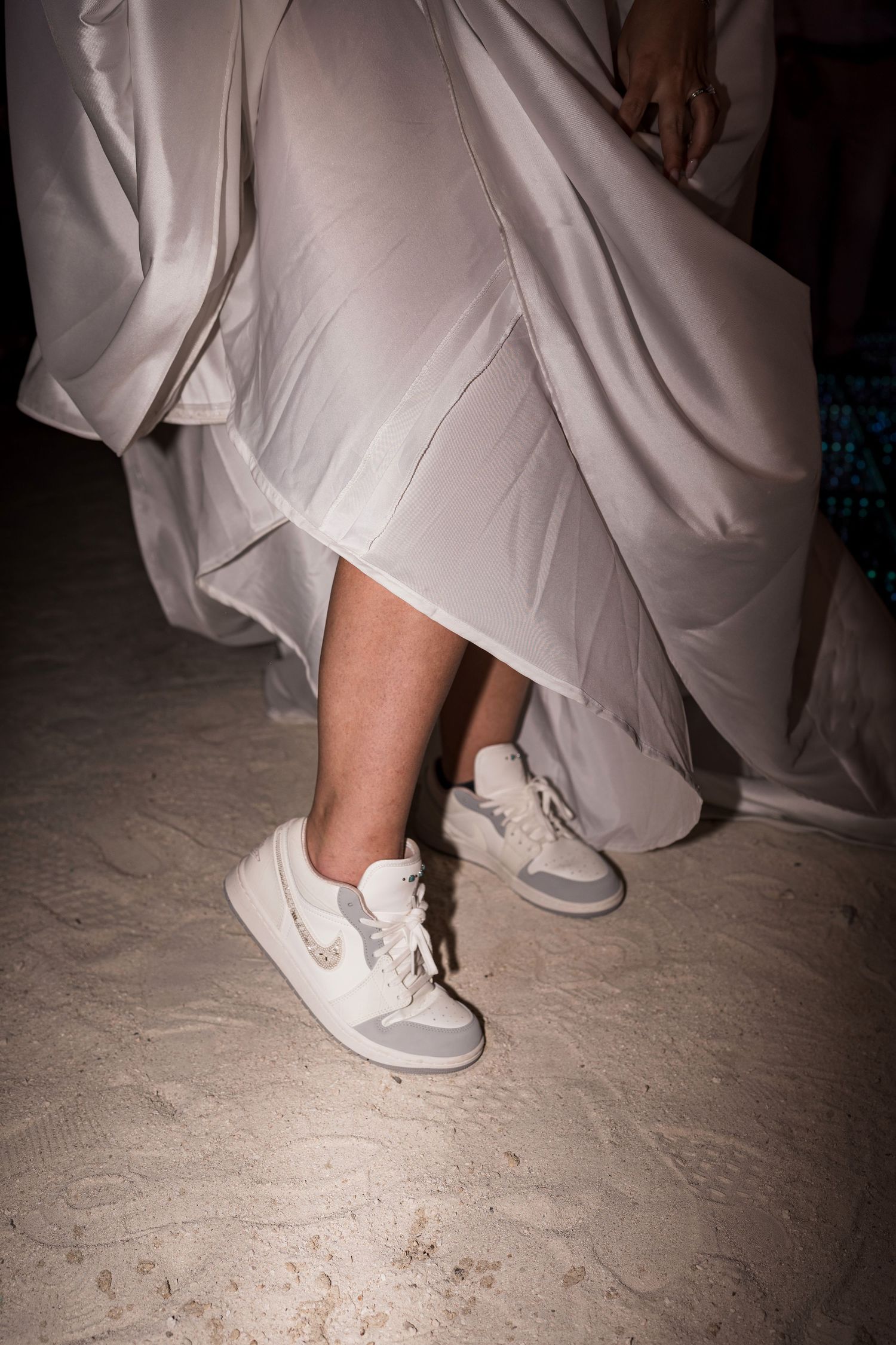 Person wearing white Nike sneakers under a flowing beige satin dress, standing on sandy ground at night.
