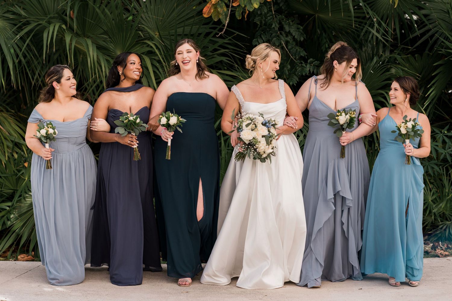 Bride in white gown stands with five bridesmaids in mix-and-match blue and gray gowns, holding bouquets outdoors.