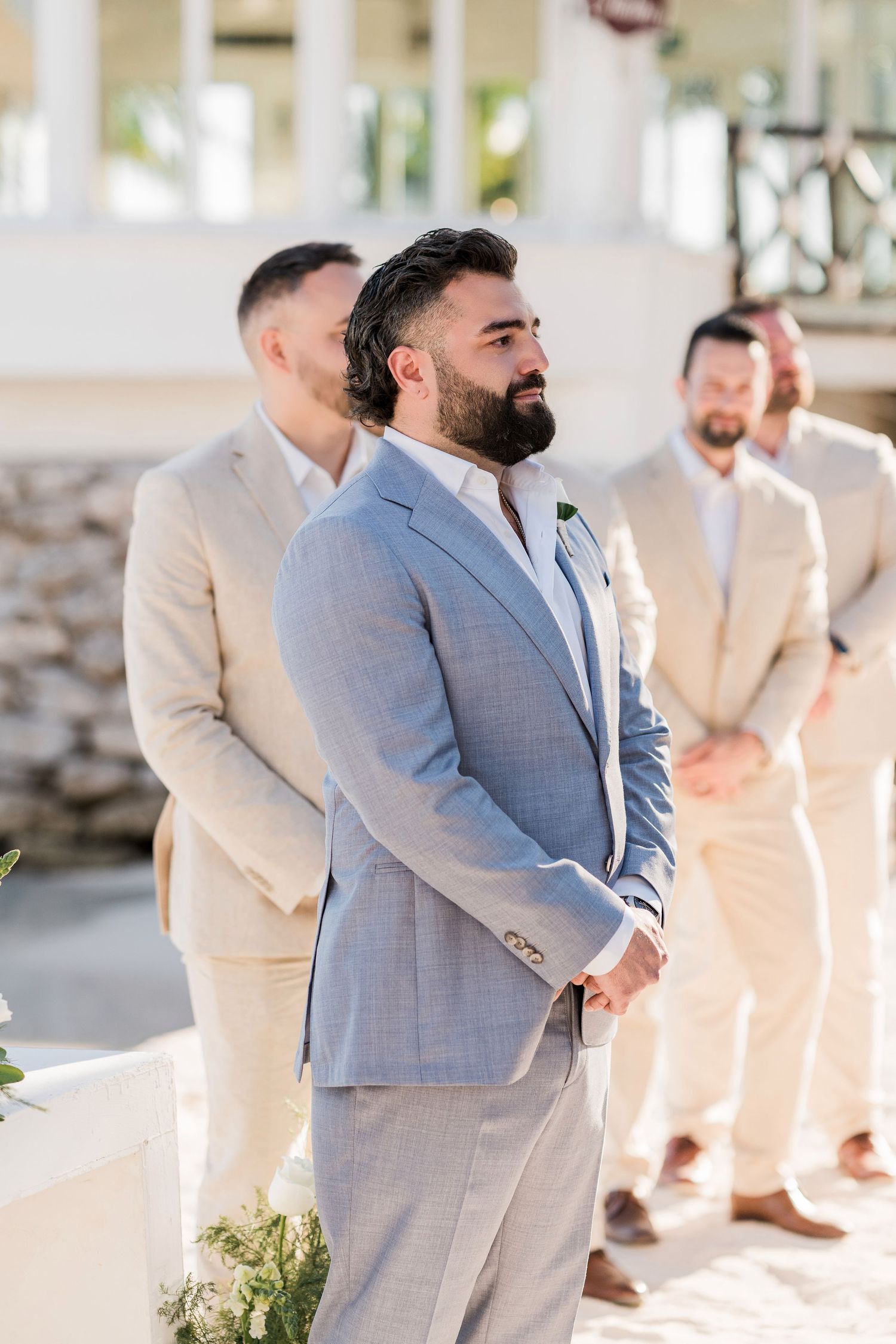 Groom in light blue suit stands at altar with groomsmen in tan suits during outdoor wedding ceremony.